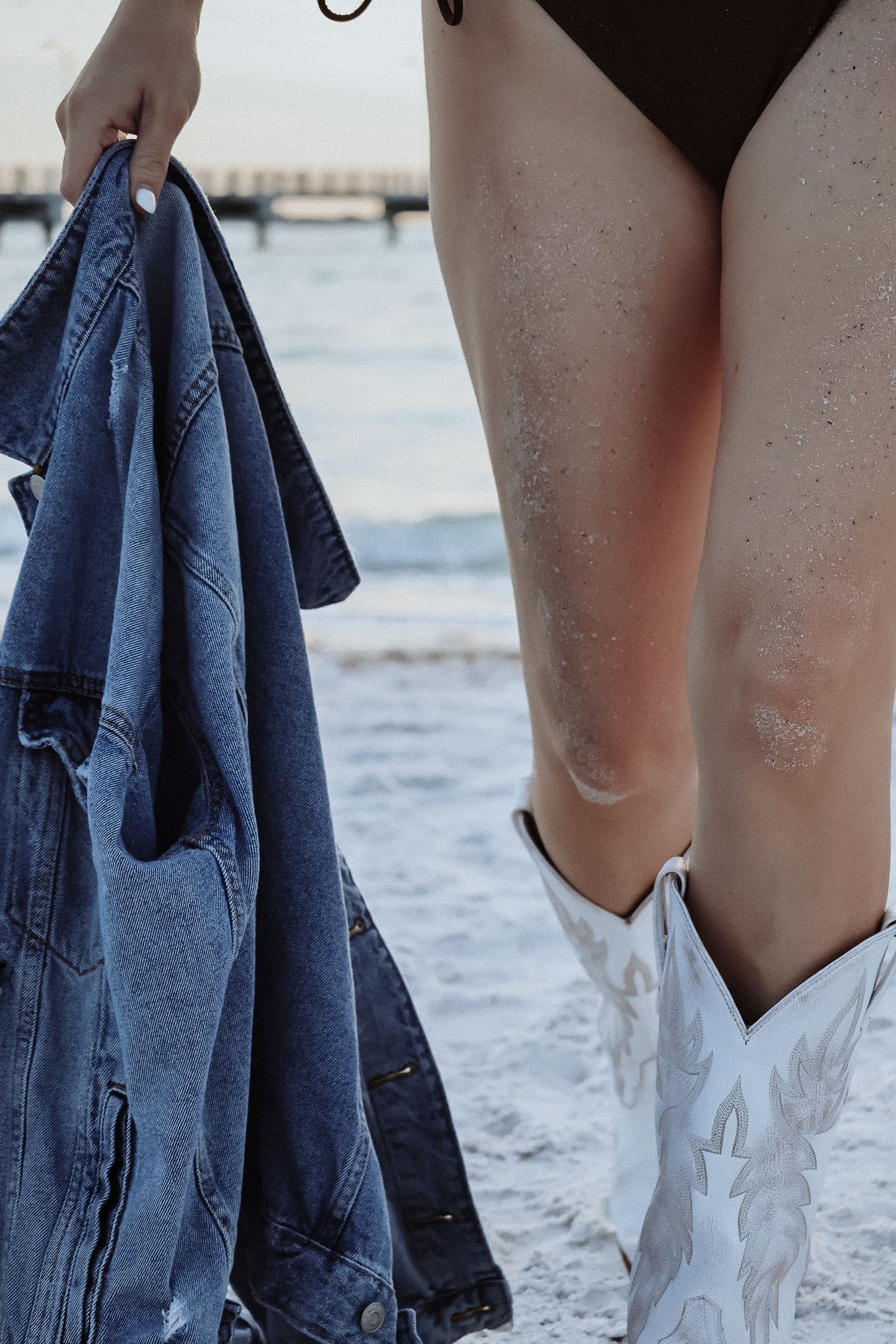 Person holding denim jacket wearing white cowboy boots at beach.