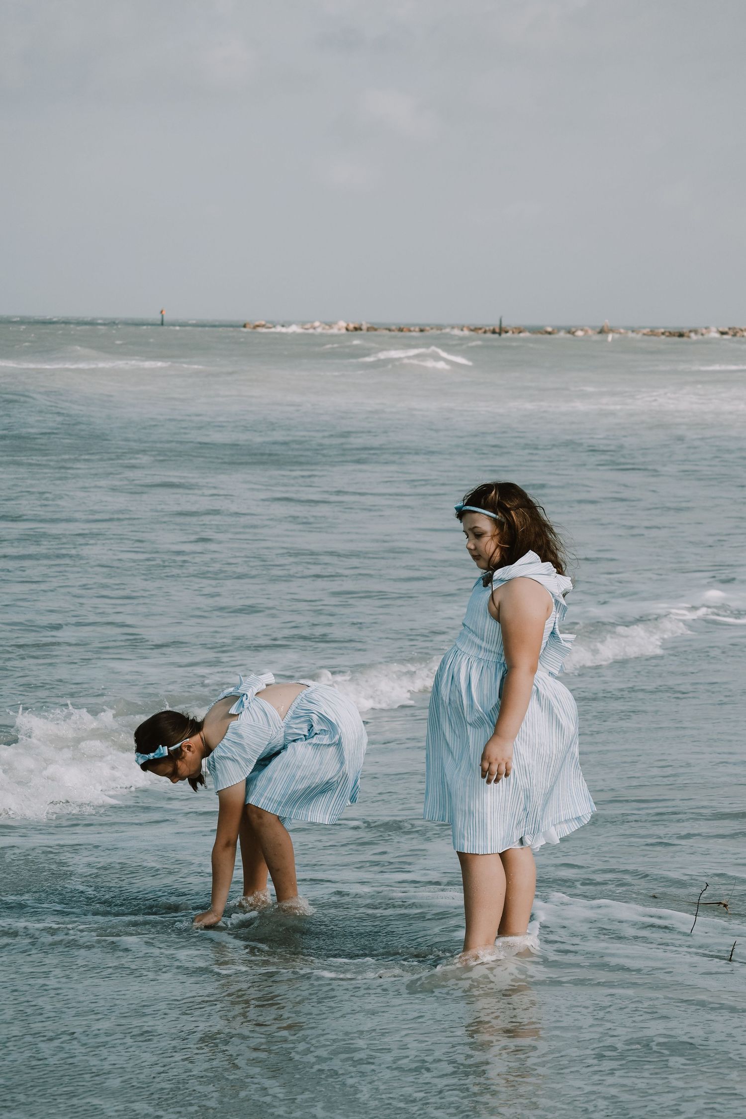 Two people in light dresses playing in the ocean waves on a cloudy beach day.