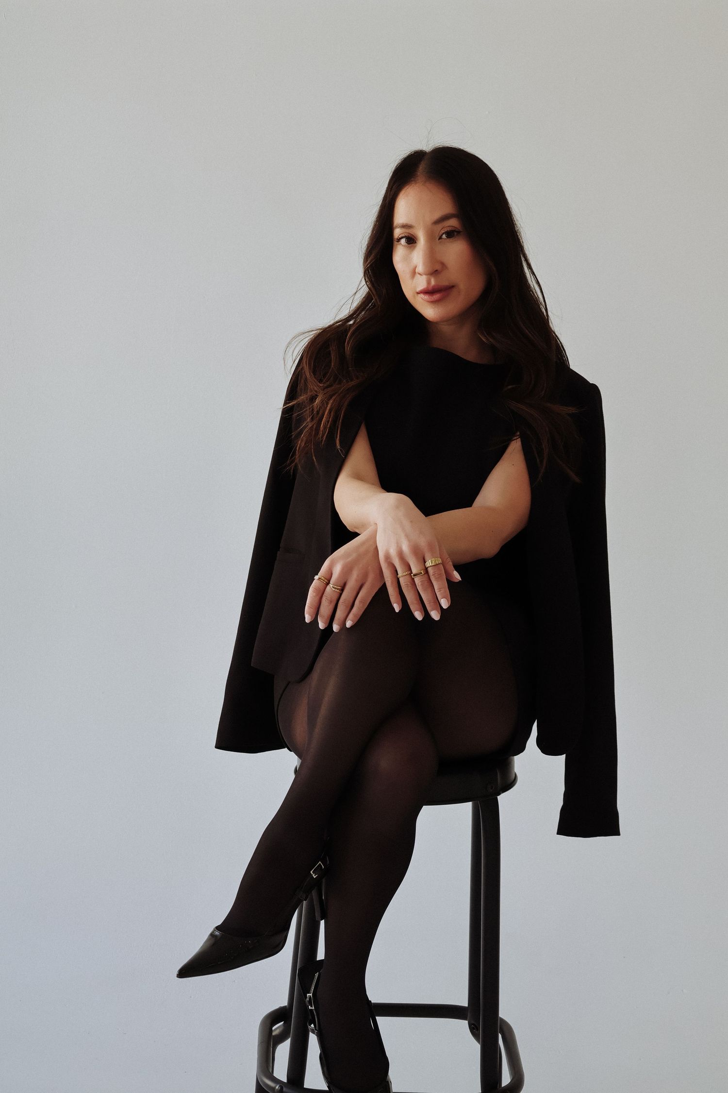 Elegant portrait of woman in black blazer sitting on stool against white background.