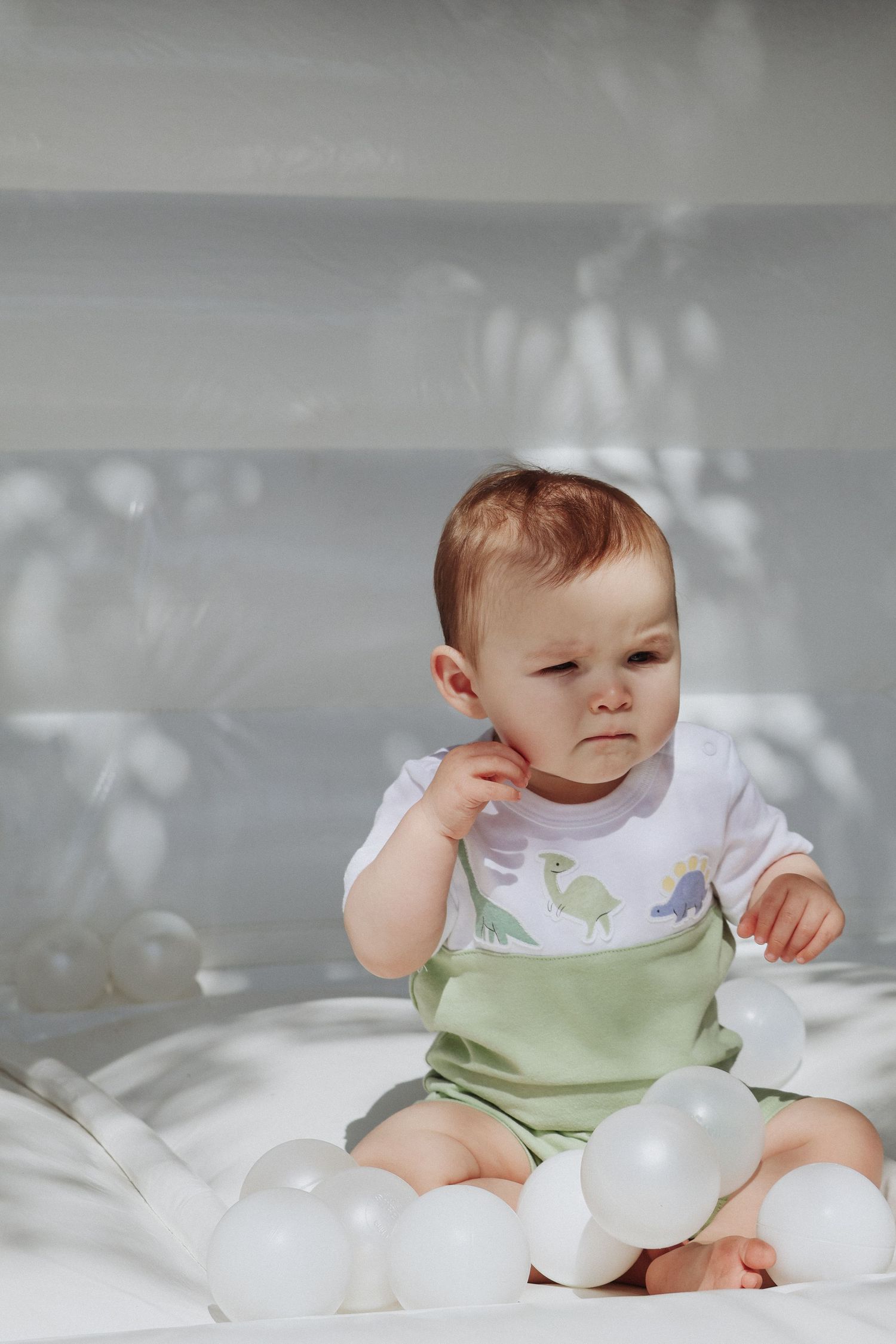 Baby in white and green outfit sitting among soft white decor.