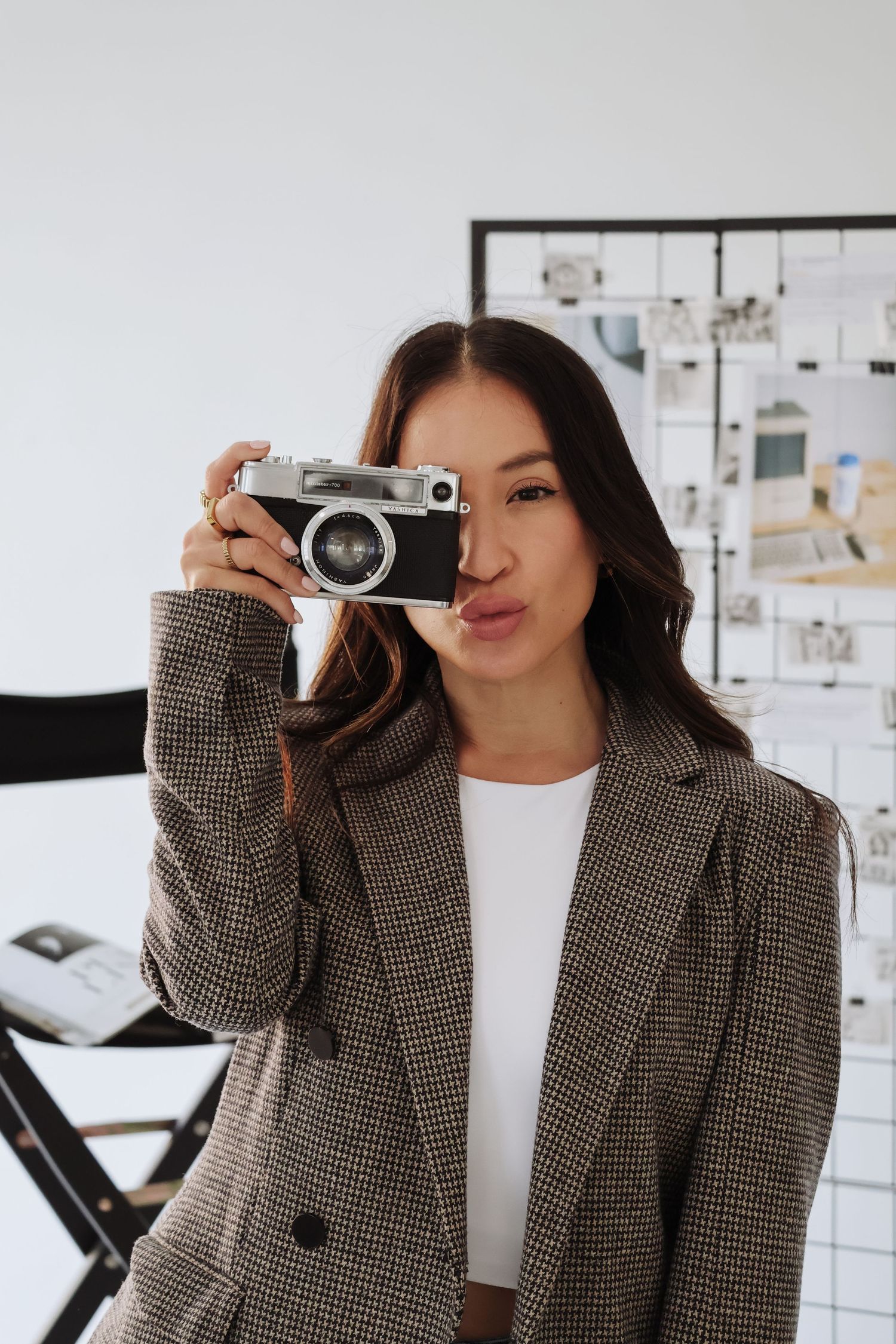 Woman in plaid blazer taking photo with vintage camera against white wall.