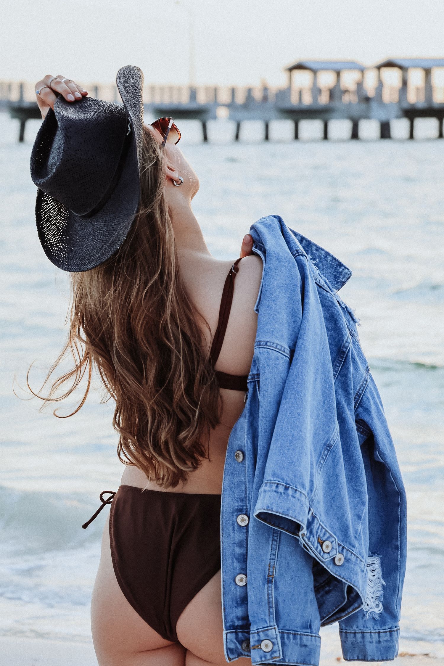 Woman in black swimsuit with denim jacket against ocean pier backdrop.