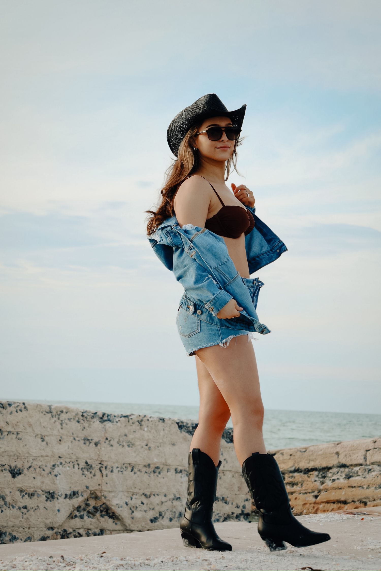 Woman in brown bikini, denim jacket and cowboy boots poses against cloudy sky at beach.