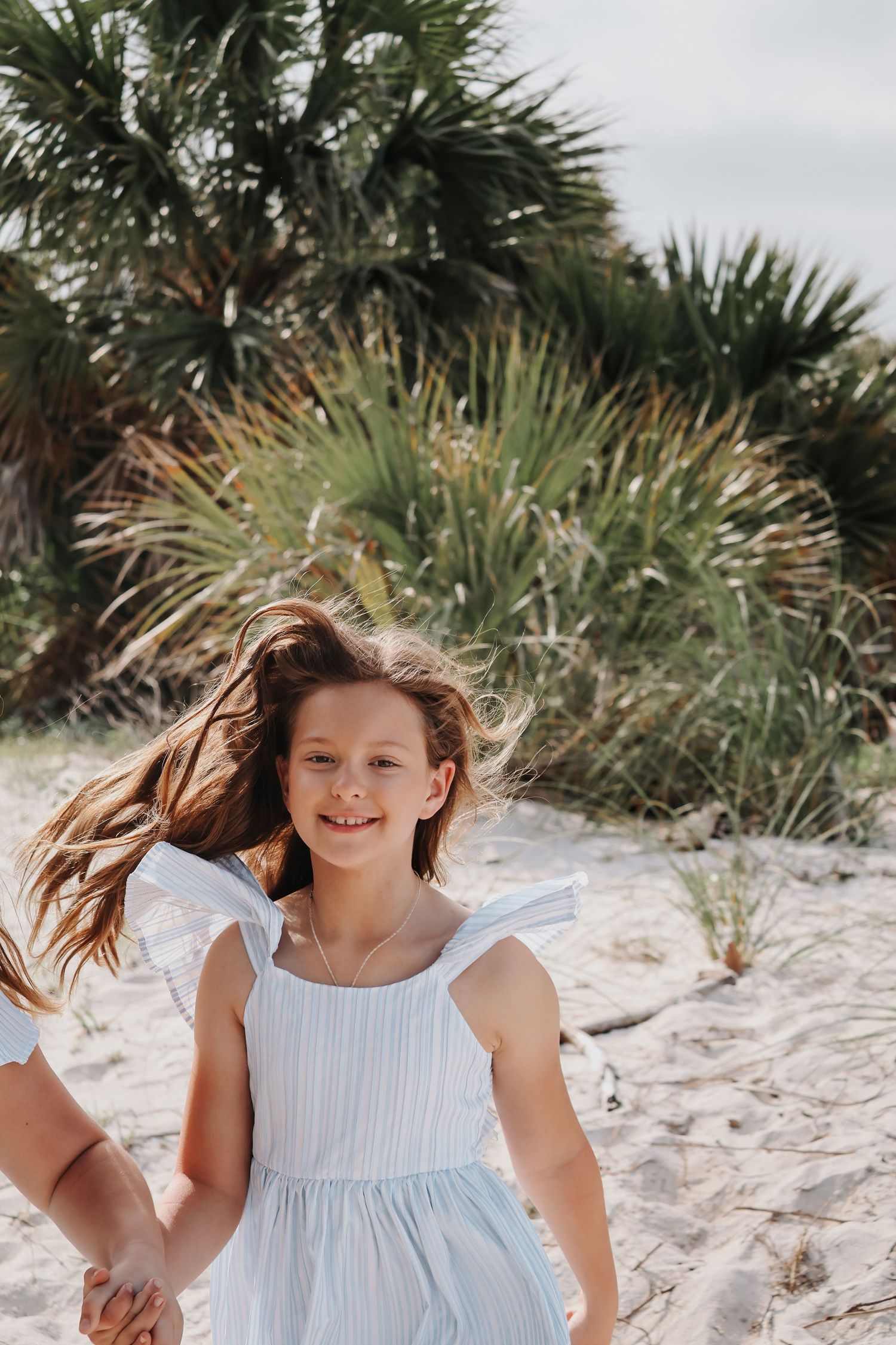 Young girl in white dress twirling and laughing on sand dunes with palm trees in background.