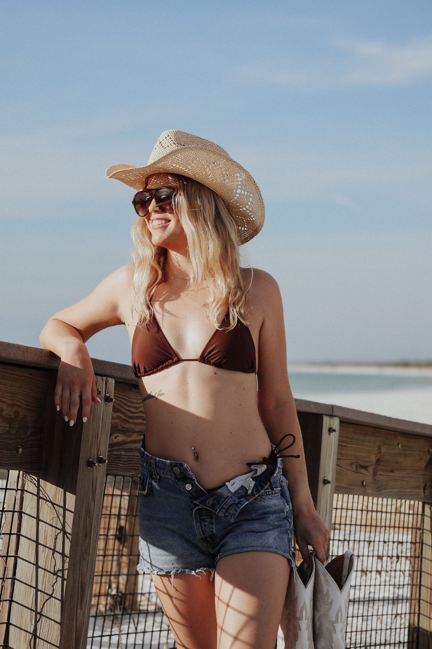 Beachwear style featuring brown bikini, denim shorts and straw cowboy hat by oceanside fence.