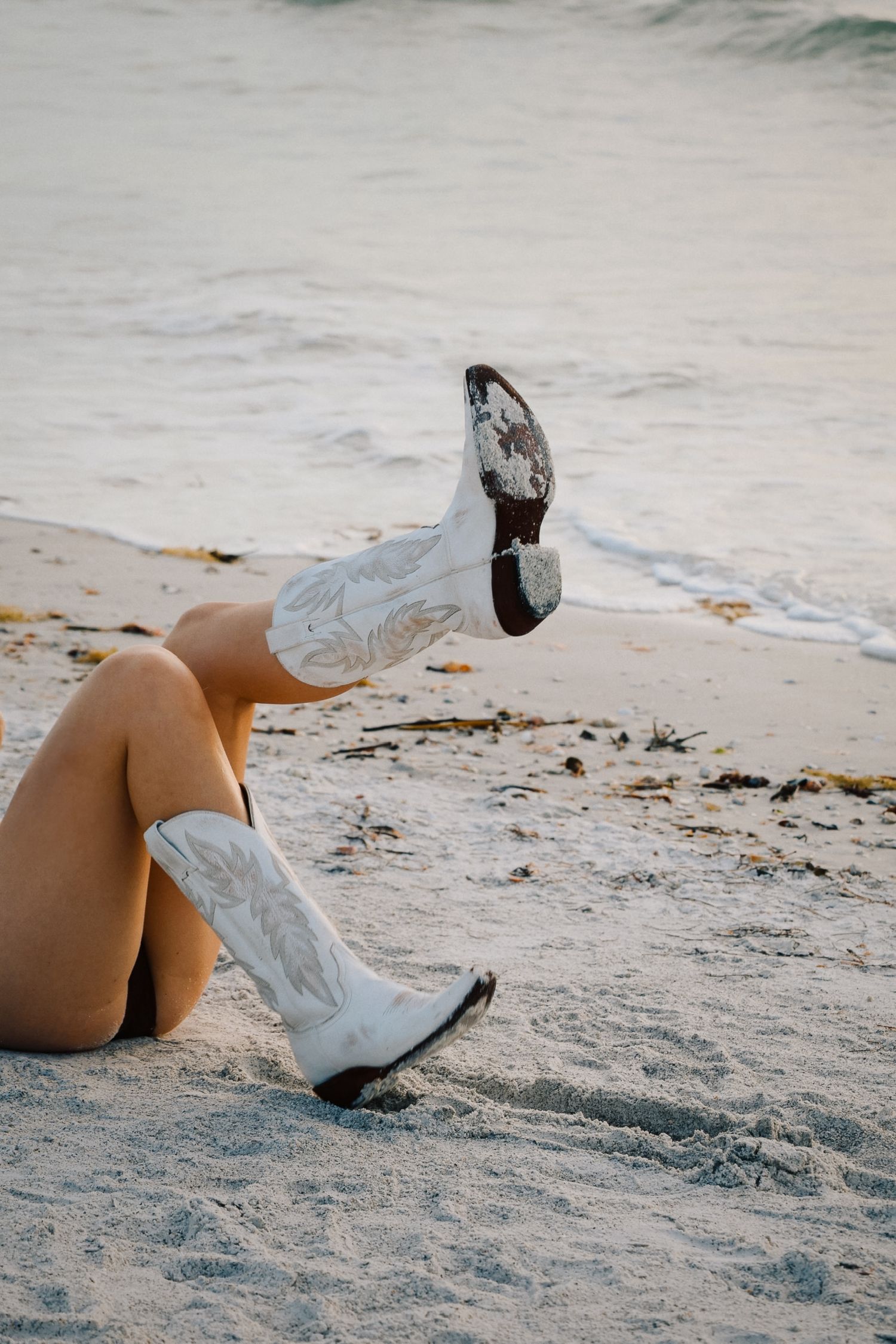 White cowboy boots against sandy beach with ocean waves in background.