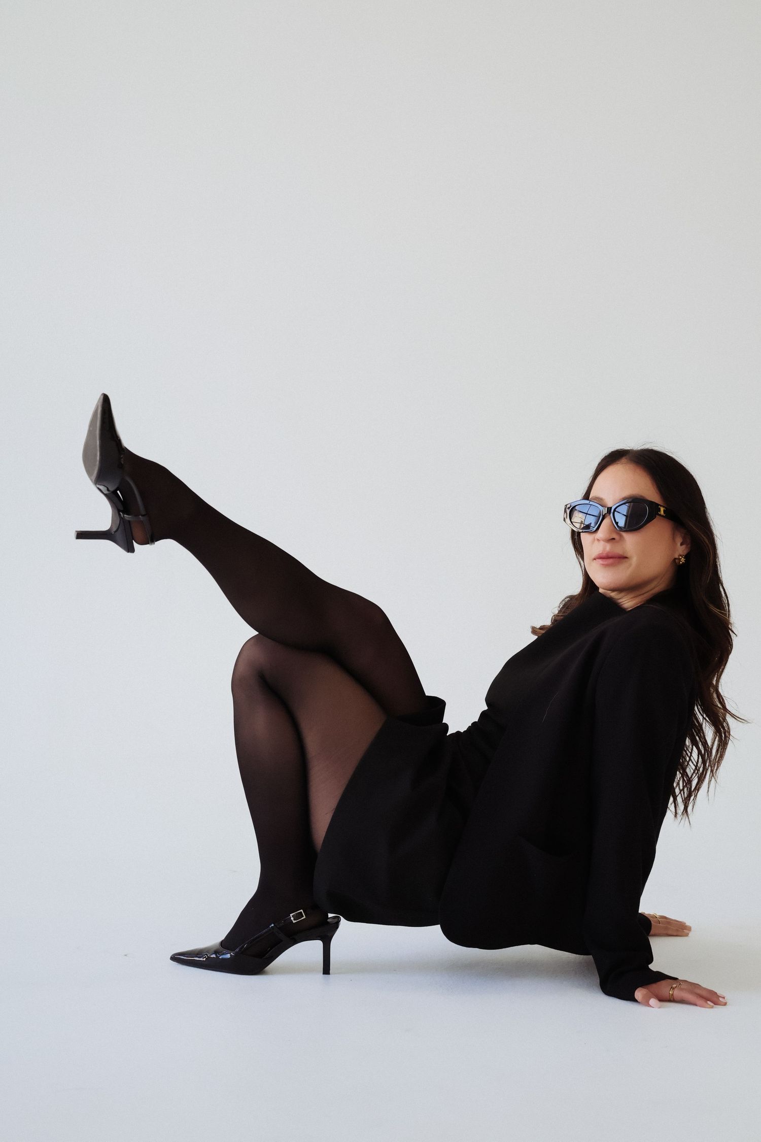 Stylish fashion pose with sunglasses and black heels against white backdrop.