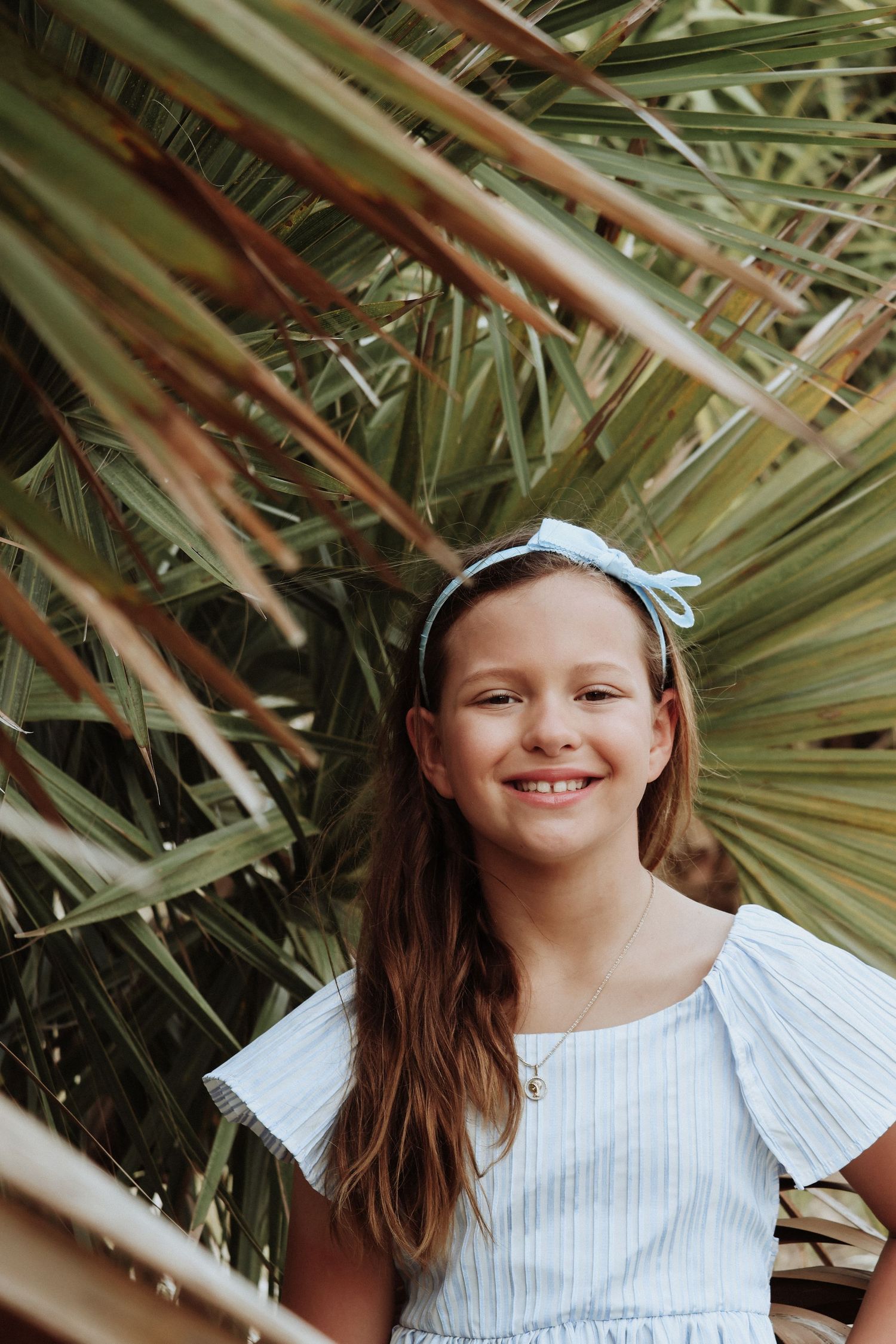 Girl smiling among tropical palm fronds wearing white dress and hair bow.