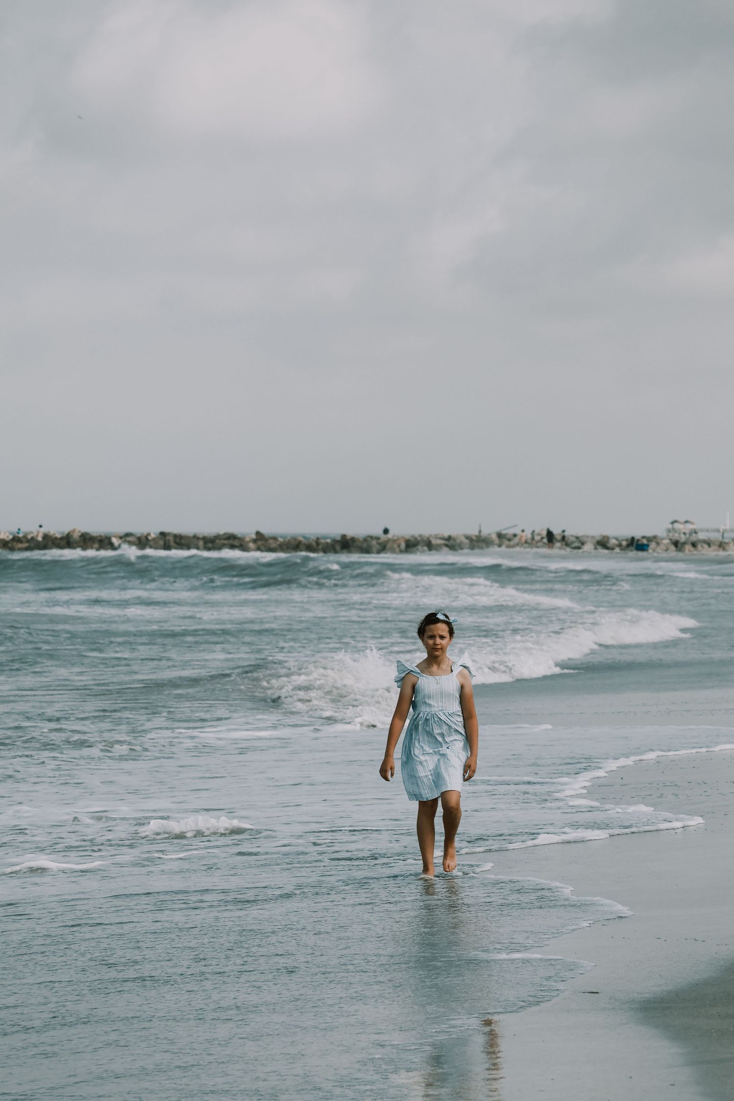 Person in light blue dress walks alone along the shoreline with waves and coastal buildings in background.
