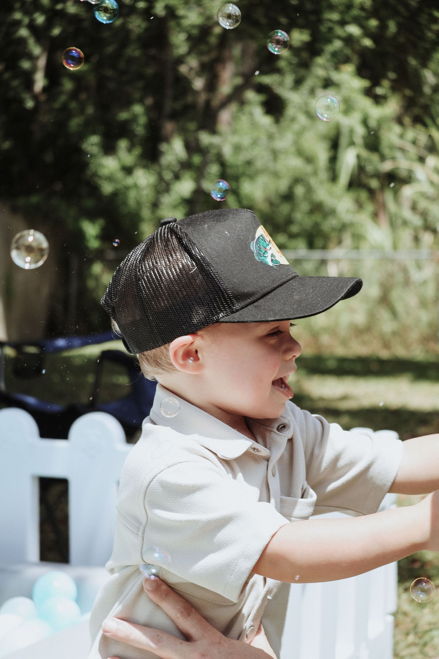 Young child in baseball cap reaching for soap bubbles in outdoor setting.