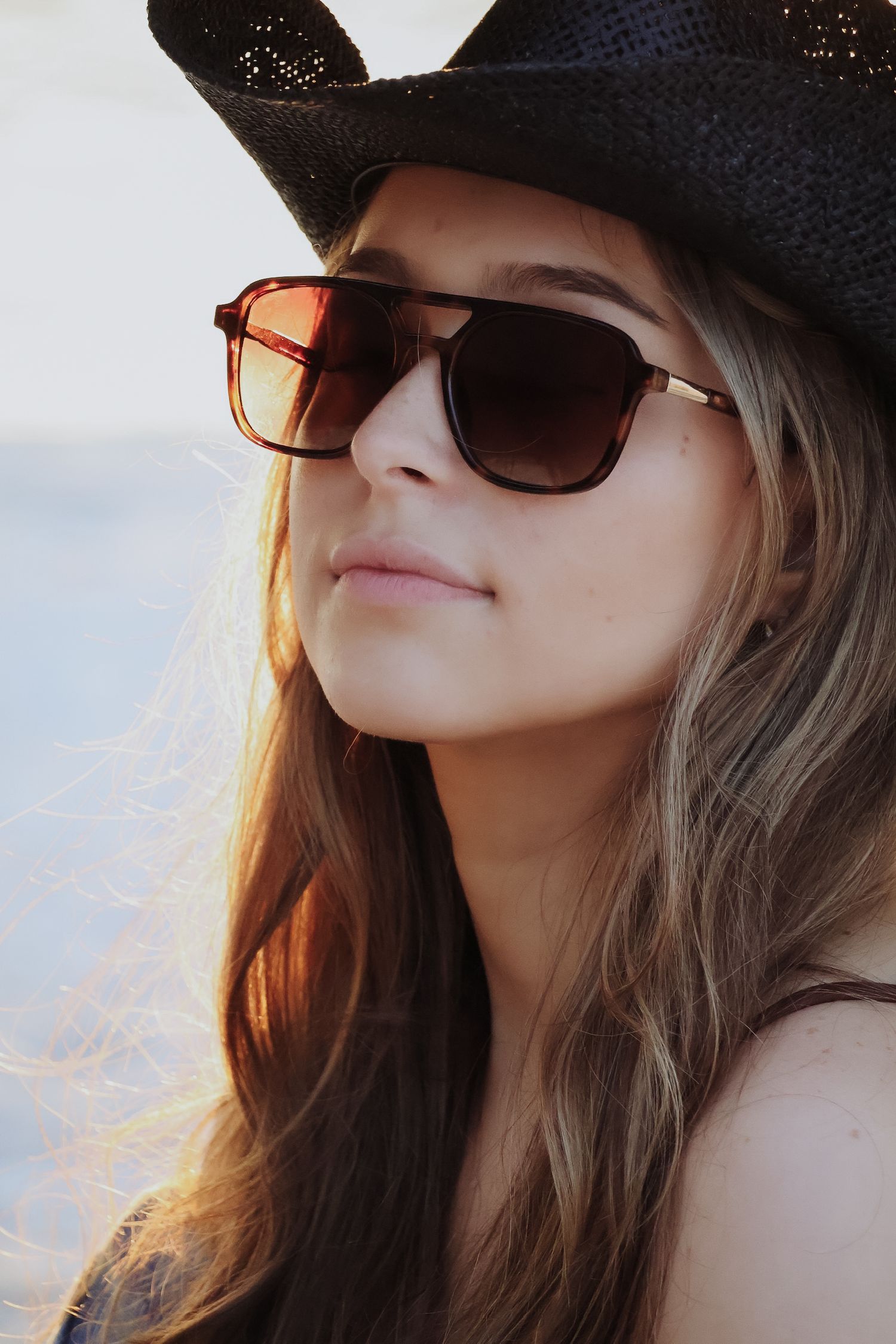 Close-up portrait of woman wearing black cowboy hat and sunglasses at sunset.