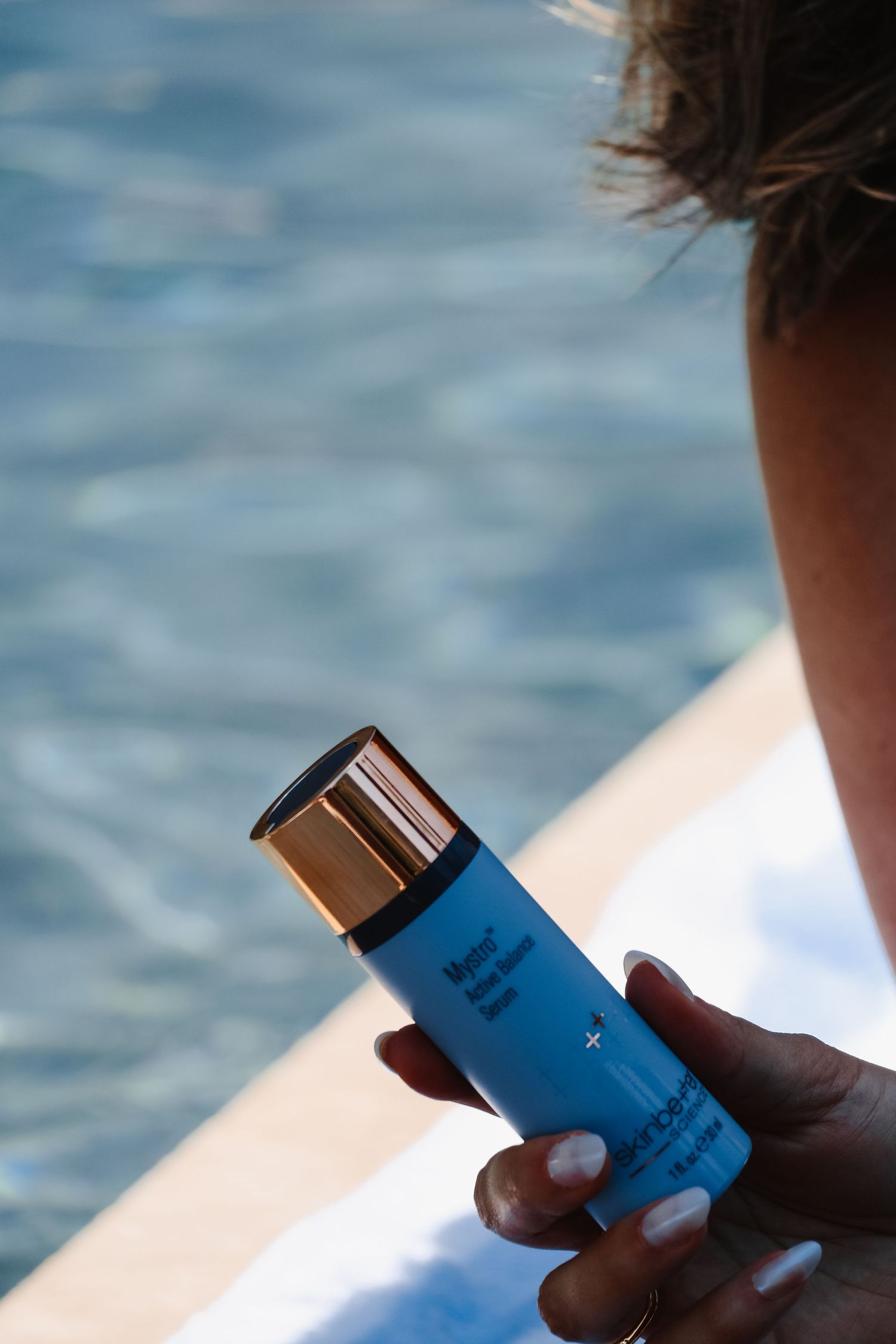 Hand holding blue skincare product bottle against blurred ocean background.