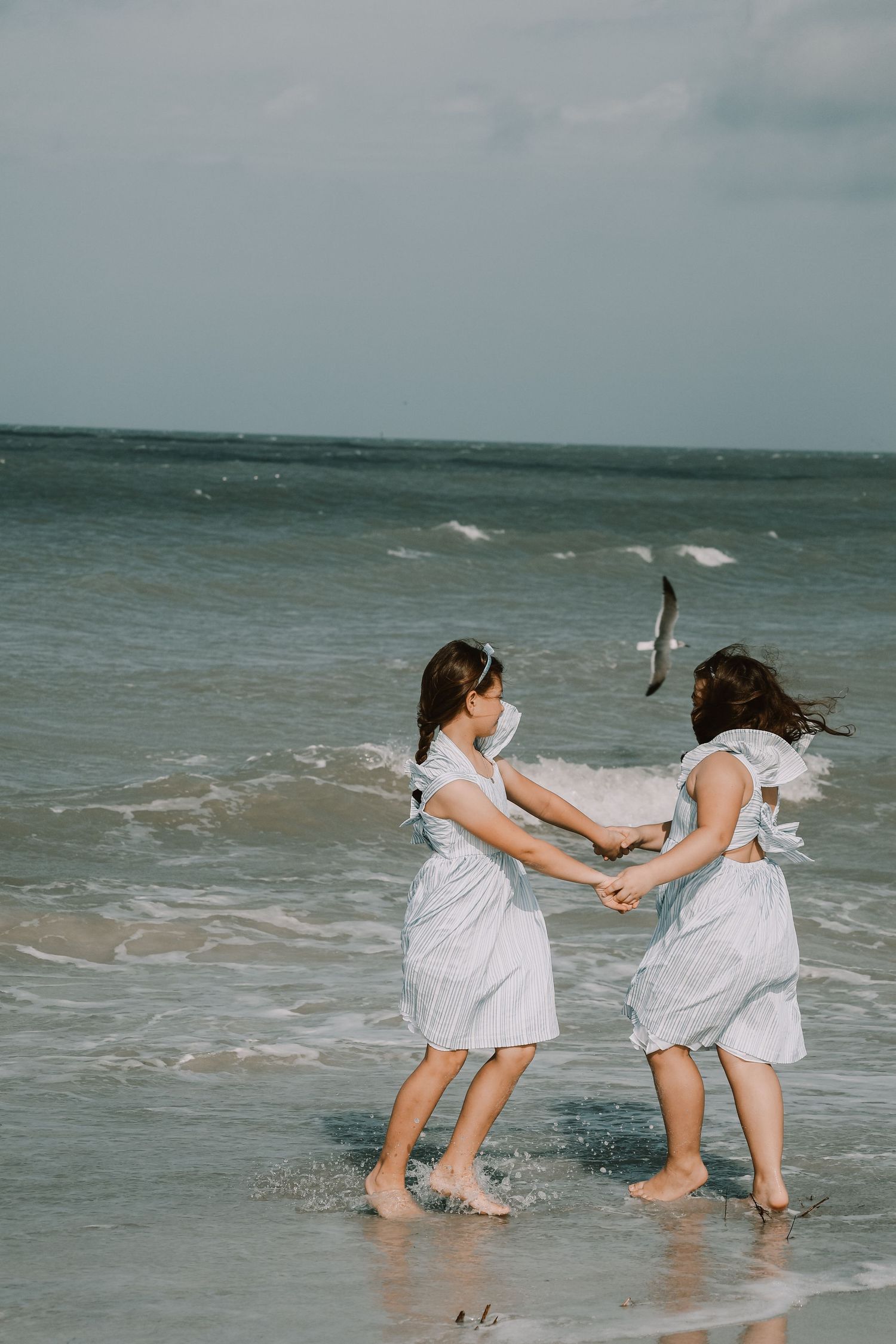 Two children playing and splashing in ocean waves while wearing matching white dresses.