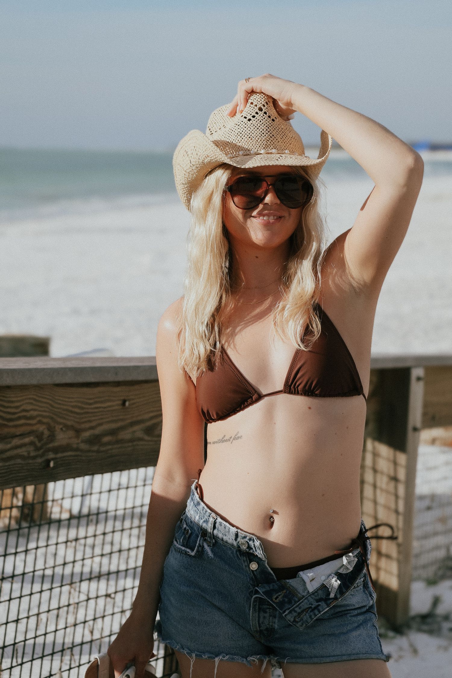 Summer beach portrait with straw hat, brown bikini and denim shorts against ocean view.
