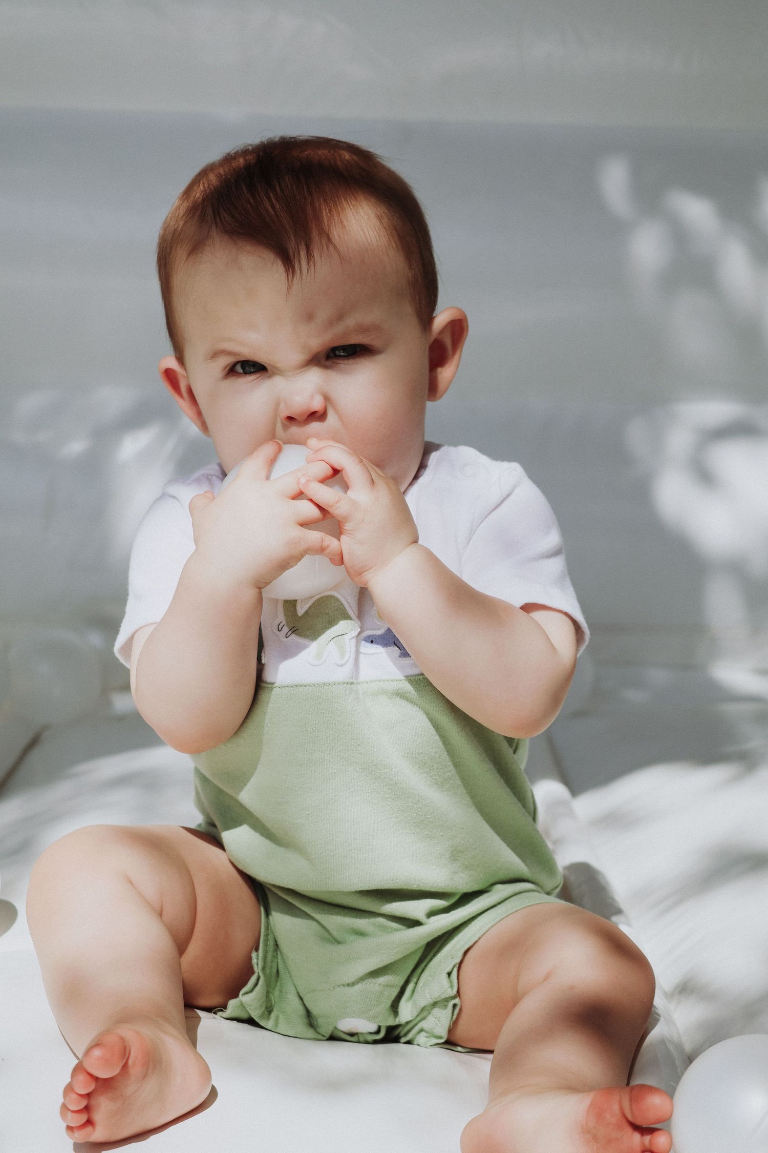 Baby in green outfit touching face while sitting on white surface.