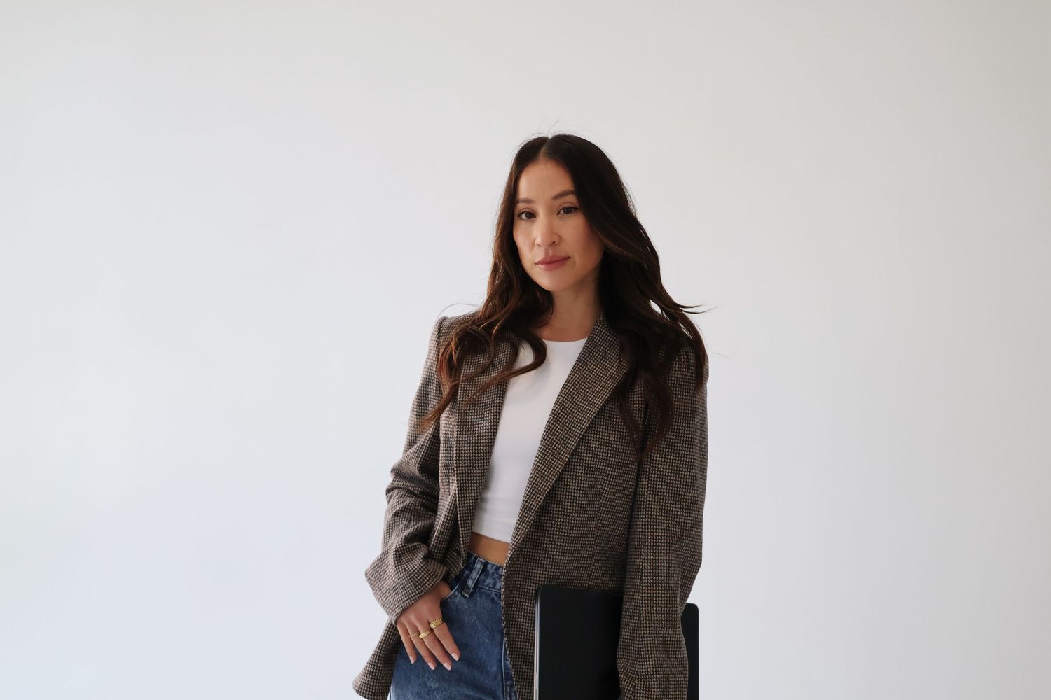 Professional woman in brown blazer and jeans posing against white wall.
