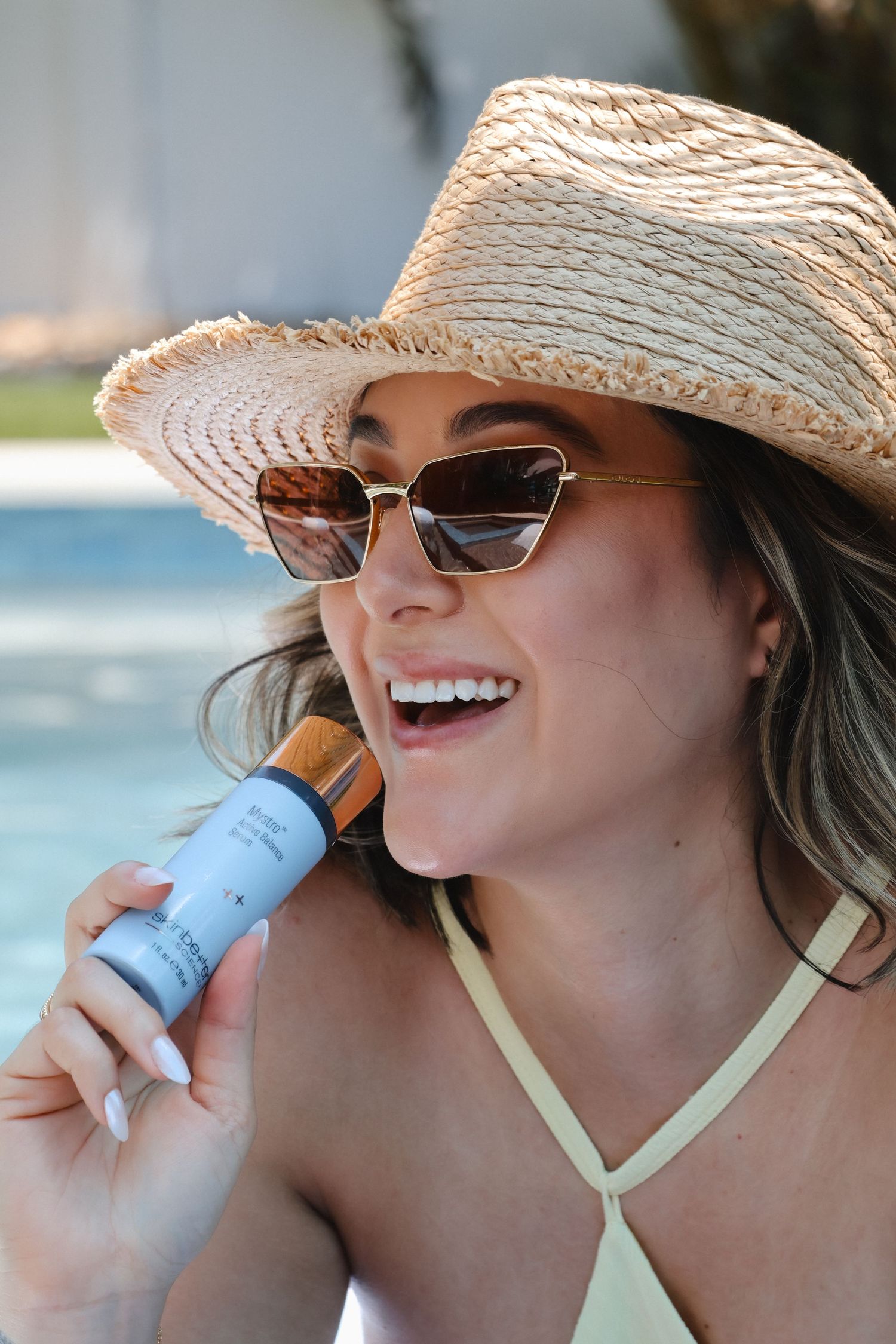 Woman in straw hat and swimsuit applying sunscreen by pool on sunny day.