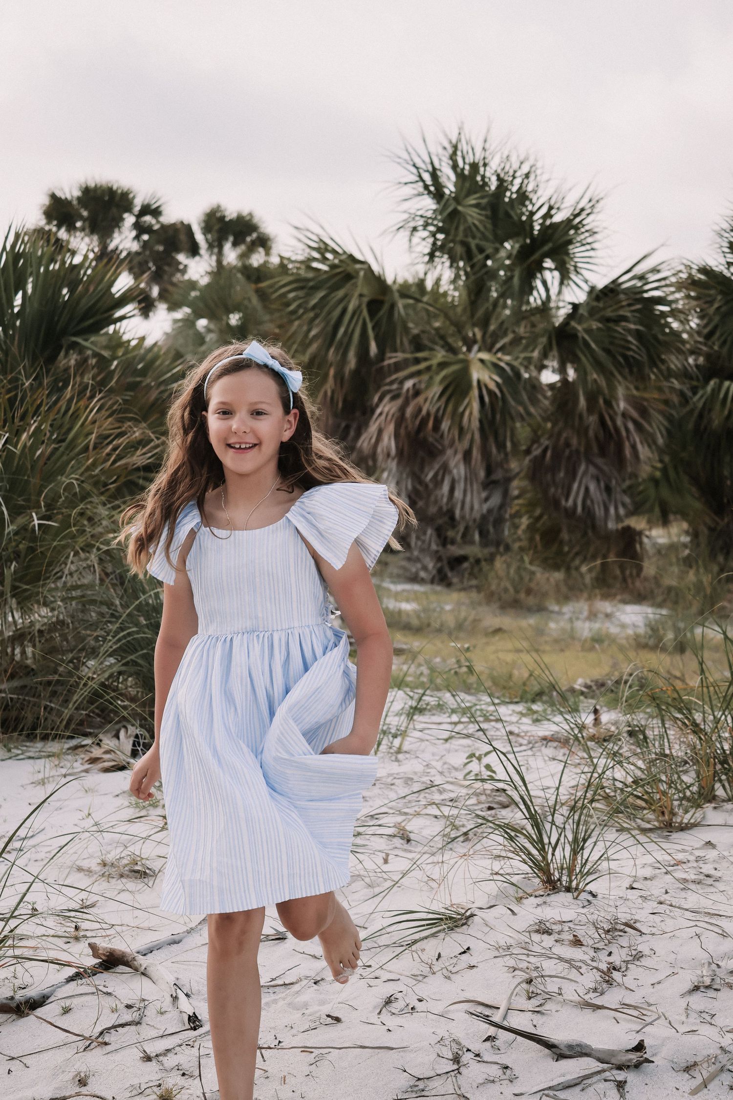 Young girl in white dress twirling on sandy beach with palm trees in background.