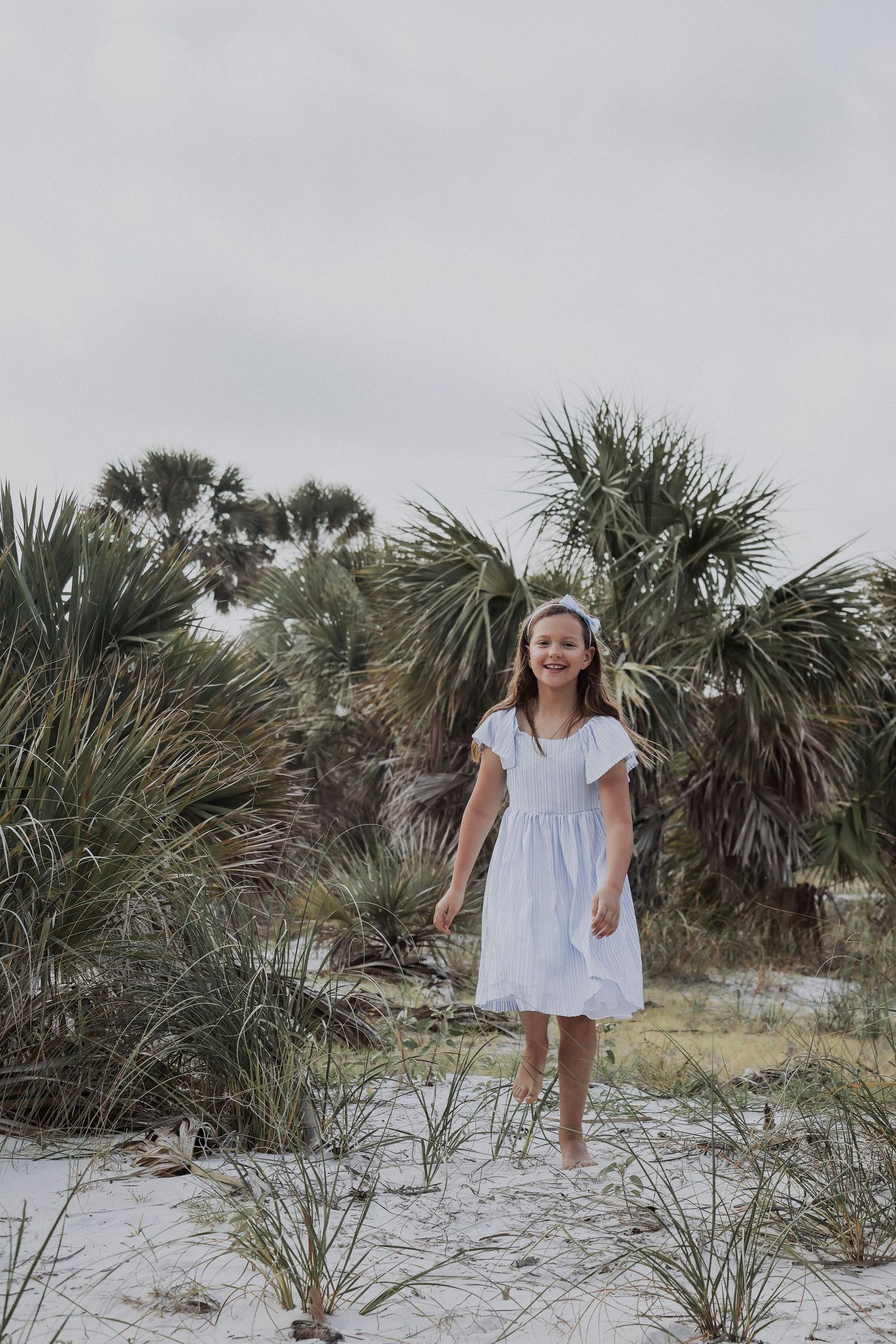 Person in white dress standing among palm trees on sandy beach path.