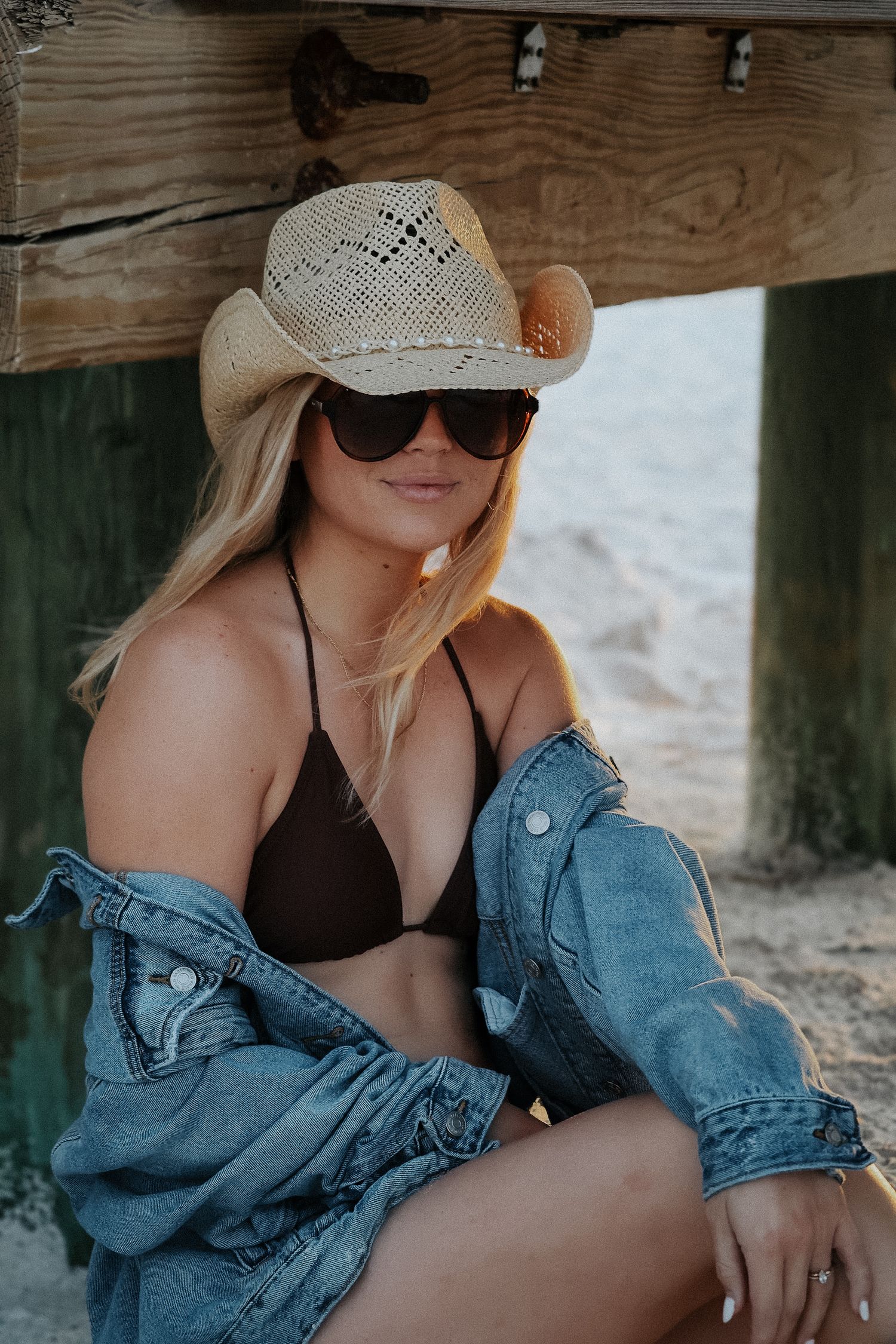 Woman in brown bikini and denim jacket poses under wooden structure wearing straw cowboy hat.