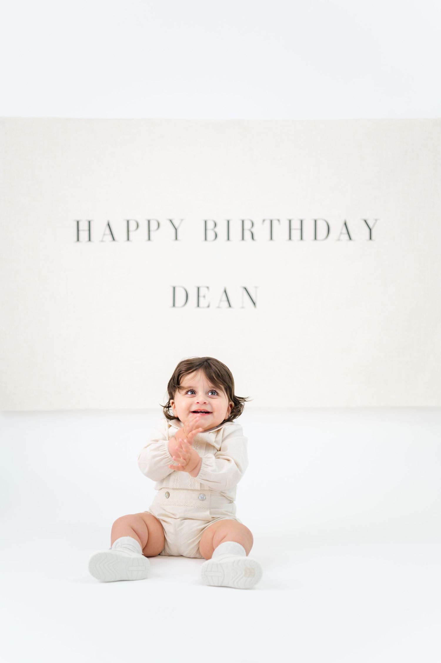 A baby in a cream outfit sits on a white floor beneath a Happy Birthday Dean message on the wall.