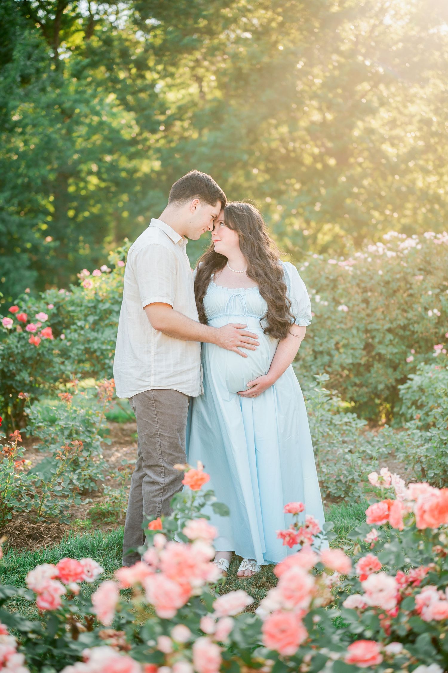 Pregnant couple sharing a tender moment in a sunlit rose garden, woman in light blue maternity gown cradling her bump.