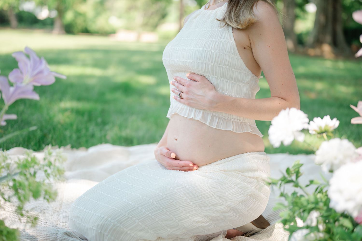 Pregnant woman in white crop top cradling baby bump outdoors among blooming flowers in a serene garden setting.