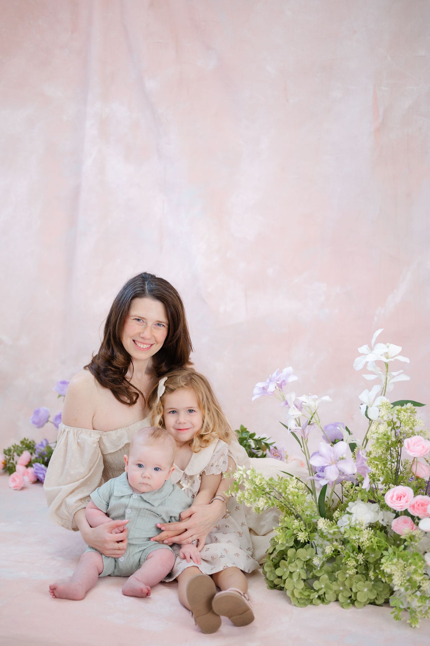 A mother sits with her two young children among pastel flowers against a soft pink backdrop in a professional portrait.