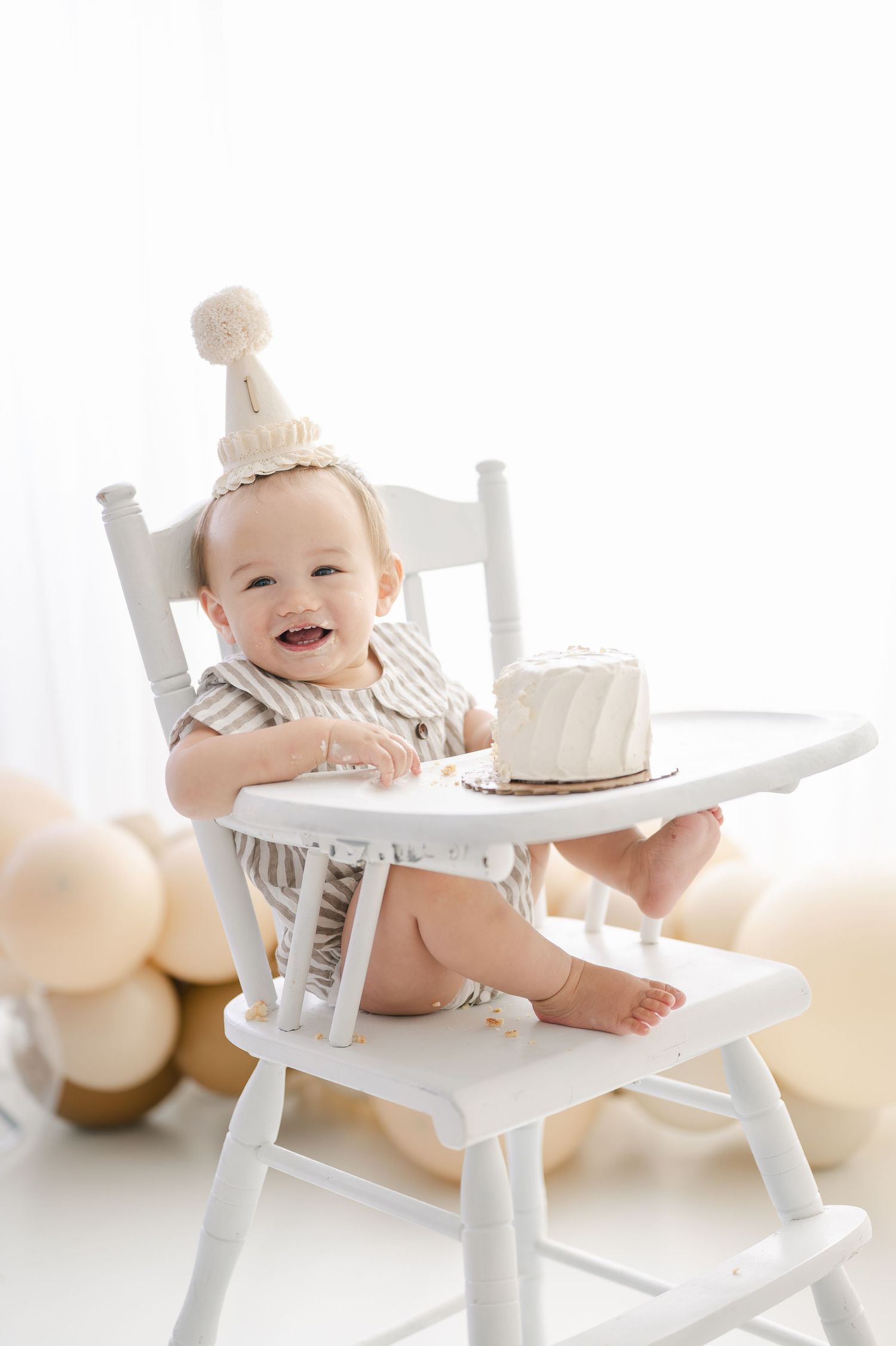 Smiling baby in white high chair wearing birthday hat with small white cake, surrounded by neutral balloons.