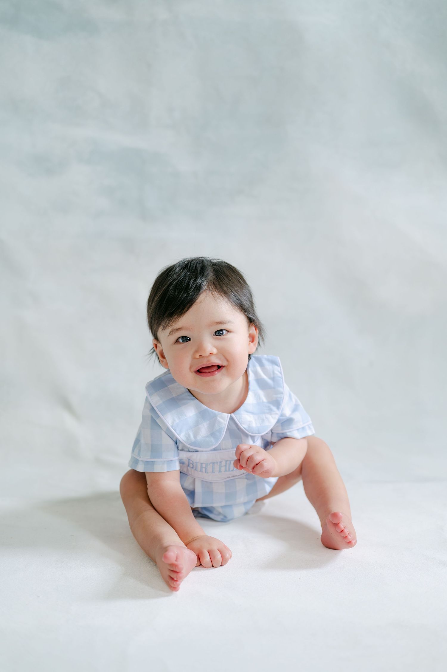Smiling baby in light blue gingham romper sitting on white background.