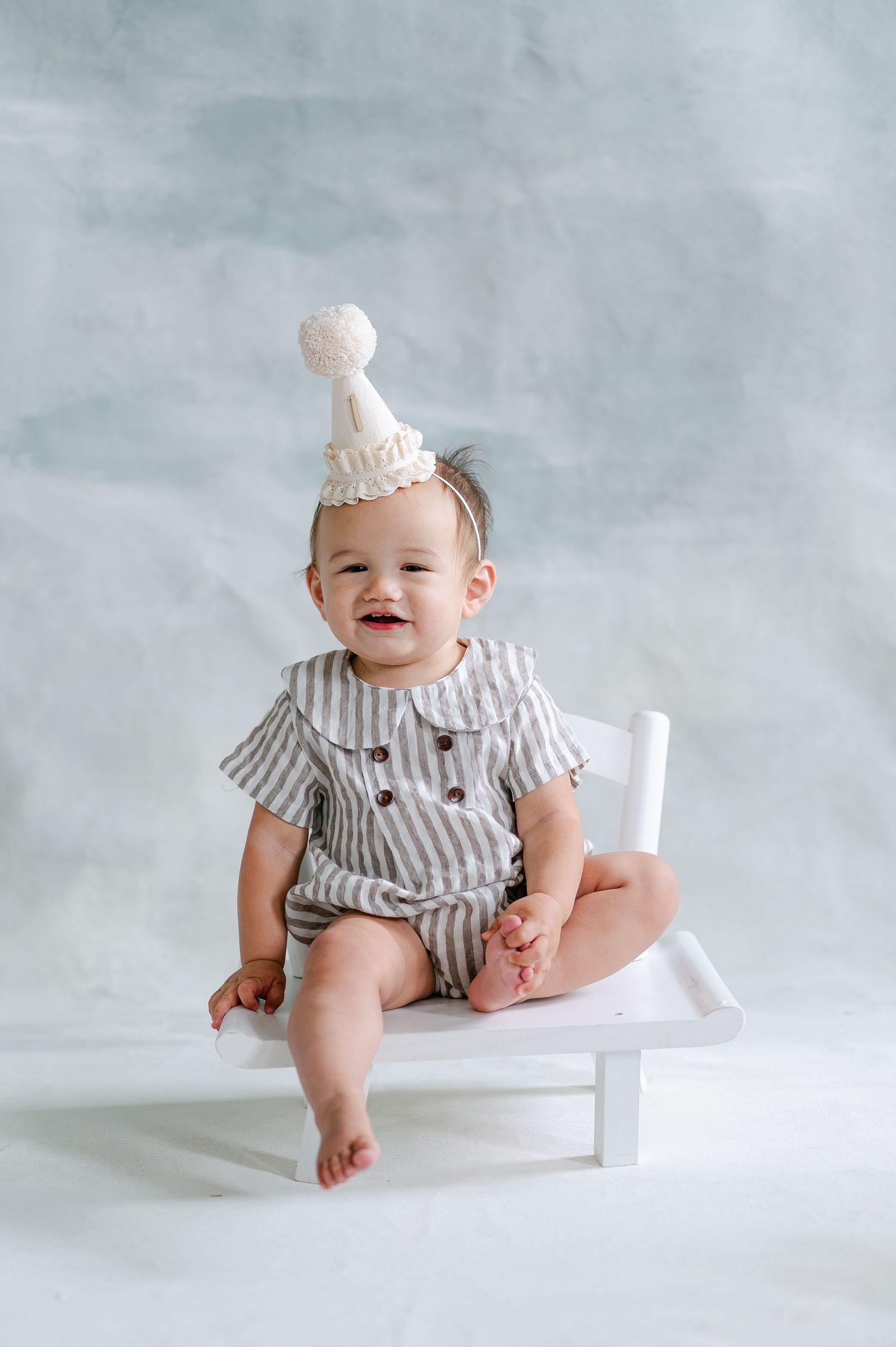 Smiling baby in striped romper and white party hat sits on small white chair against light gray backdrop.