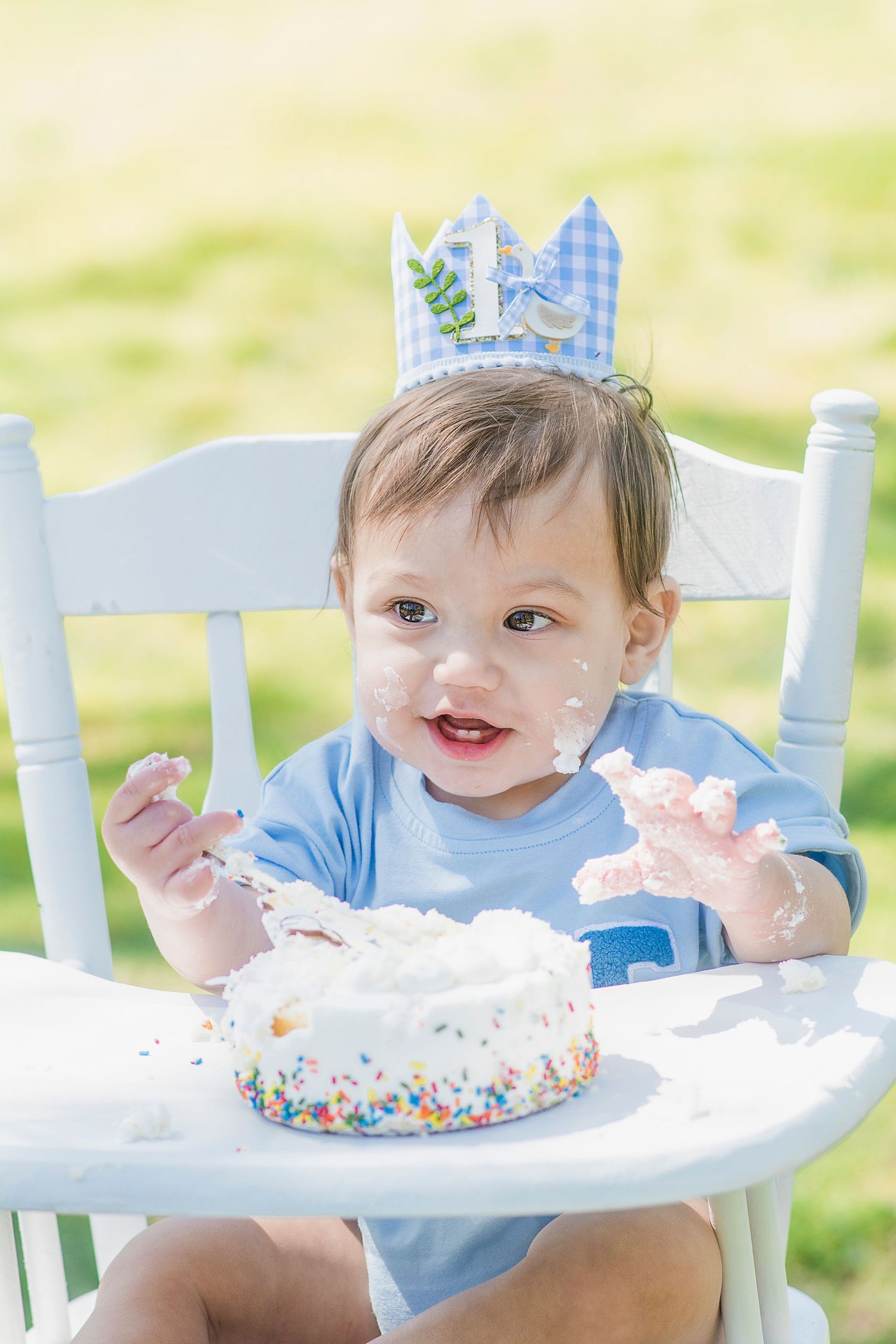 Baby gleefully plays with birthday cake in white vintage high chair during outdoor photoshoot.