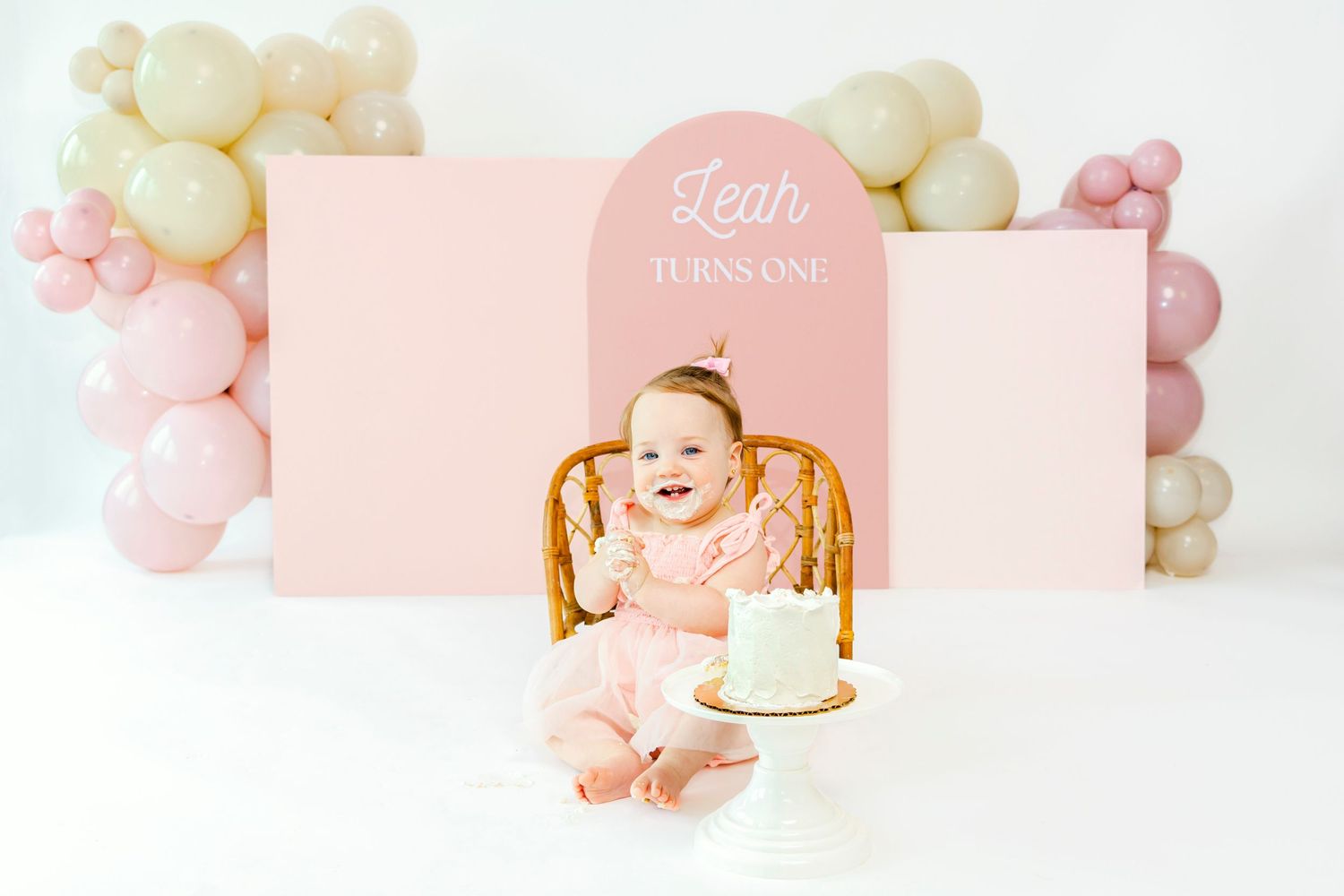 Pink and white balloon garland decorates a first birthday cake smash photography scene.