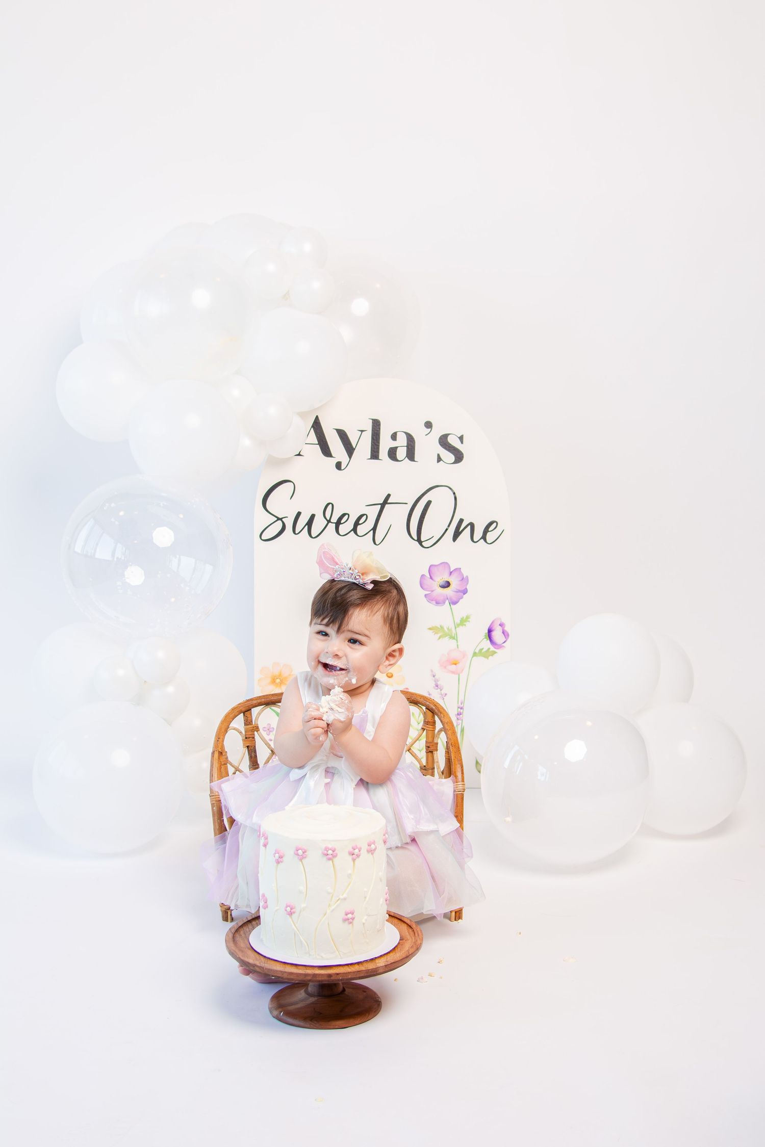 First birthday celebration setup with white cake and floral decorations against light backdrop.