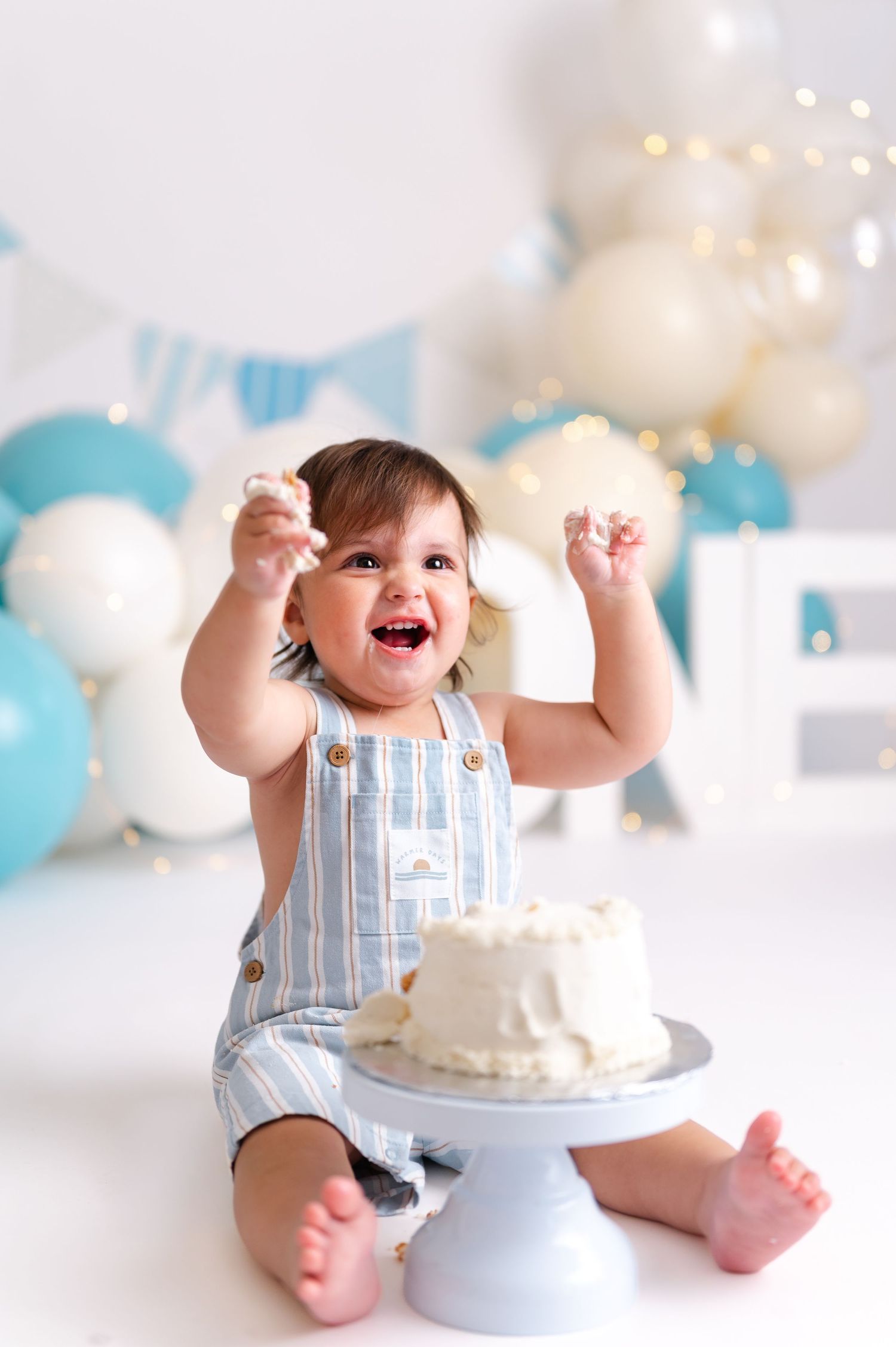 Happy baby boy in striped overalls celebrates first birthday smashing white cake surrounded by blue and white balloons.