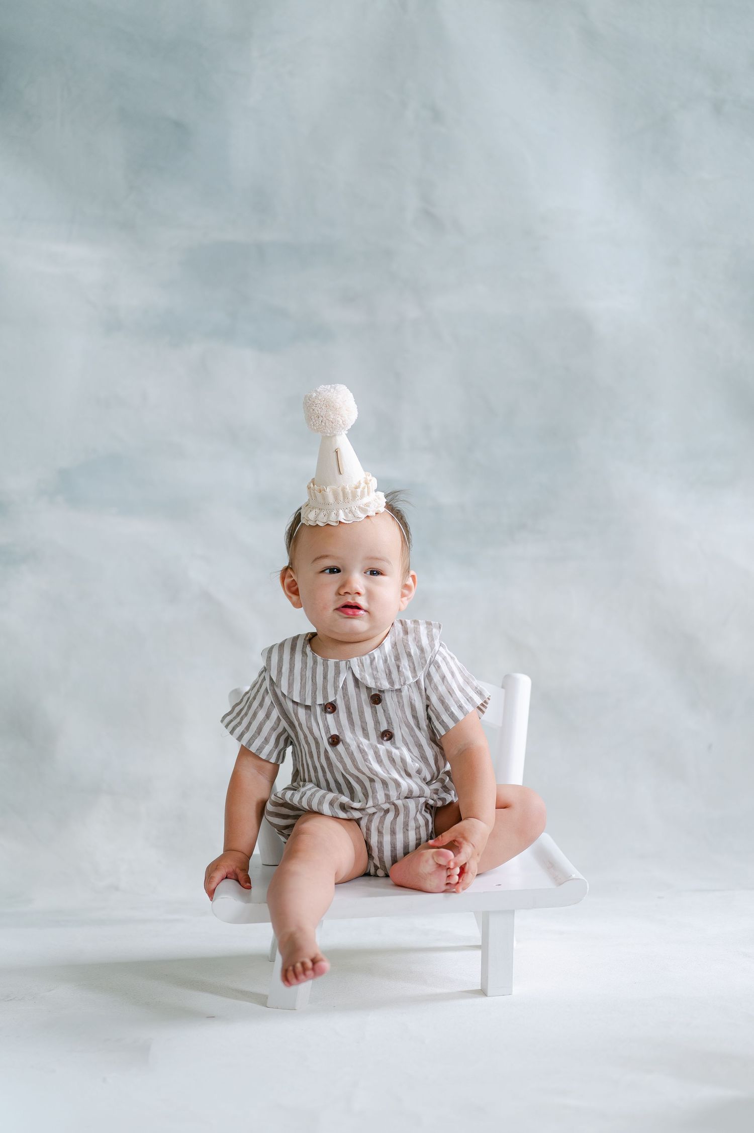 A cute baby in a striped romper and white party hat sits on a white bench against a soft gray backdrop.