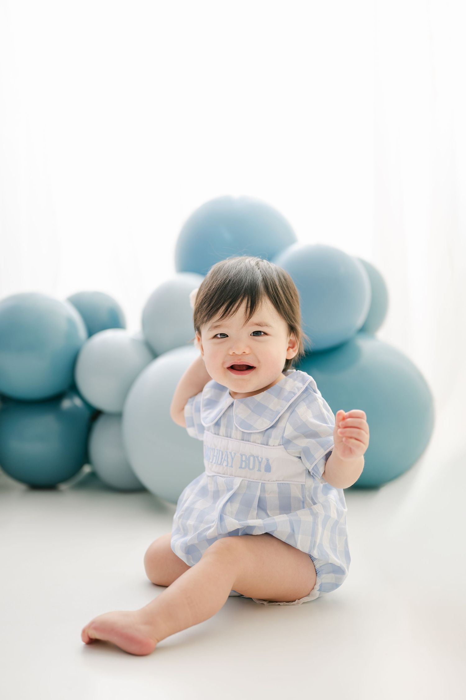 Smiling baby boy in blue striped romper sitting on white floor with blue balloon cloud backdrop.