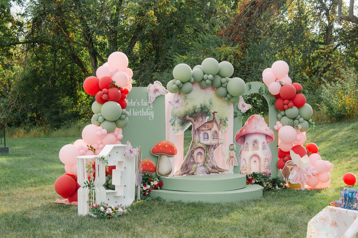 Romantic outdoor party setup with pink and red balloons, floral arrangements, and white furniture in a garden setting.