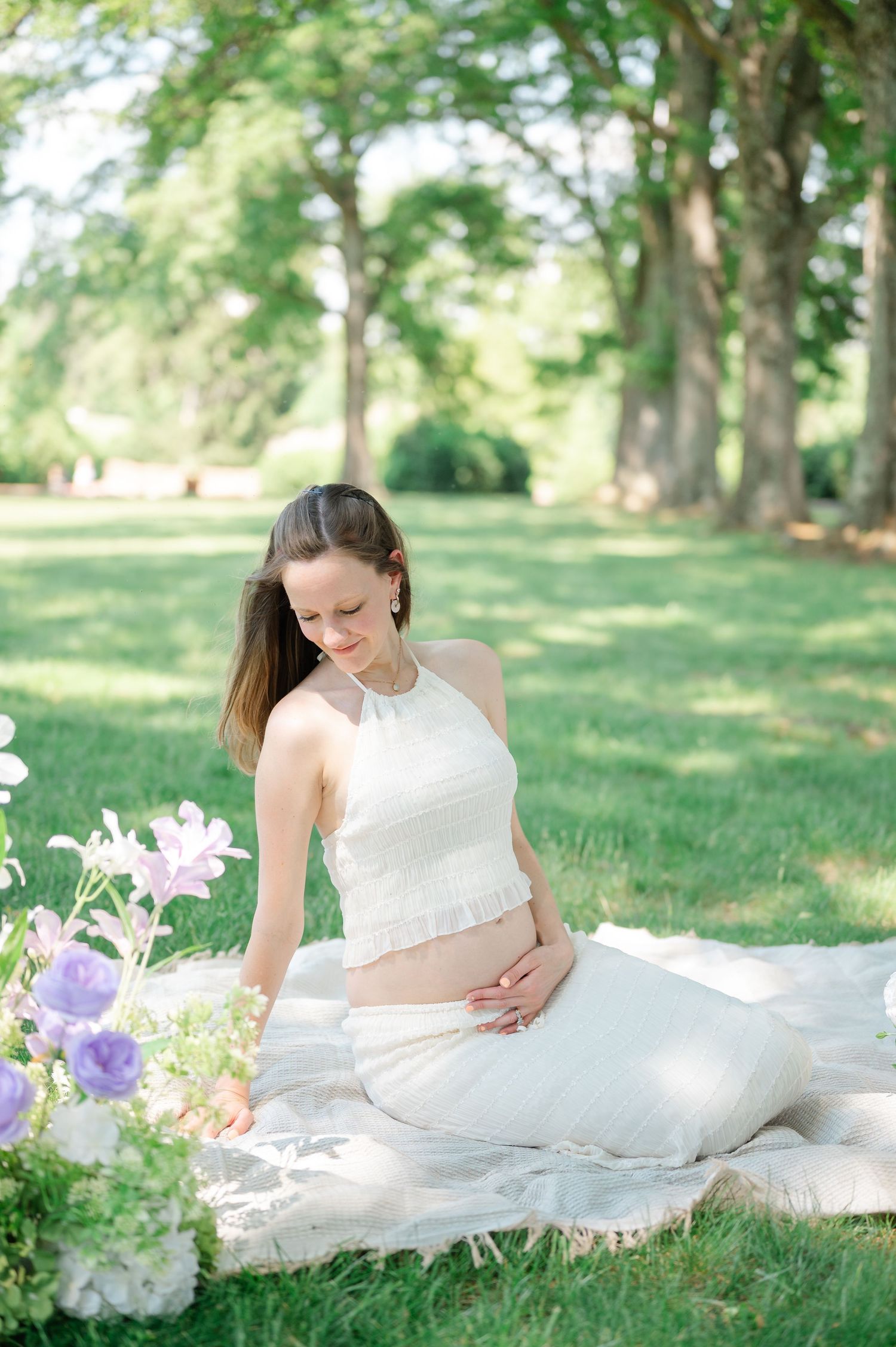 Pregnant woman in white crop top and skirt sits on blanket in sunny park, cradling baby bump near purple flowers.