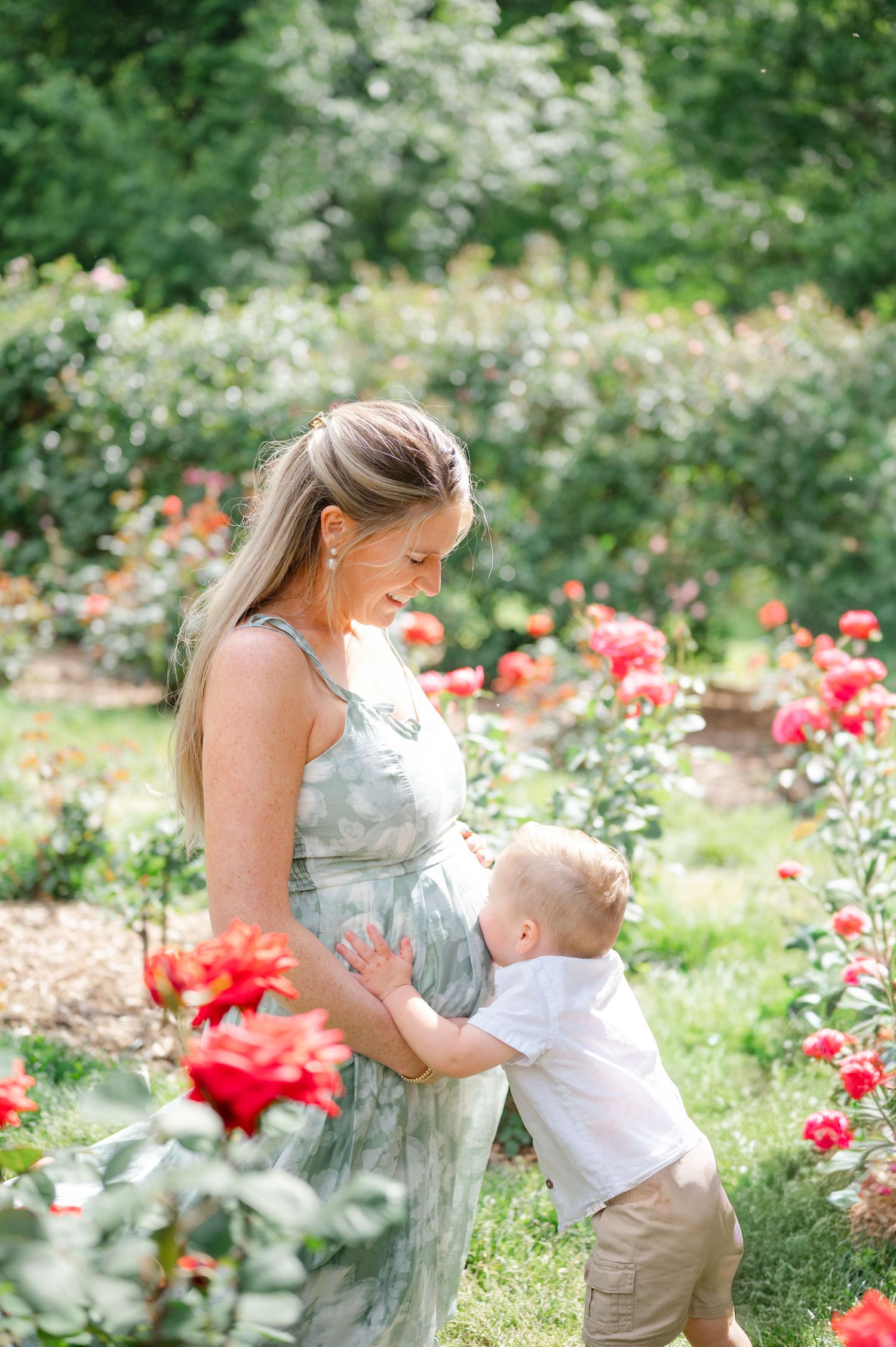 Pregnant woman in floral dress smiling at toddler touching her belly in a garden with red roses.