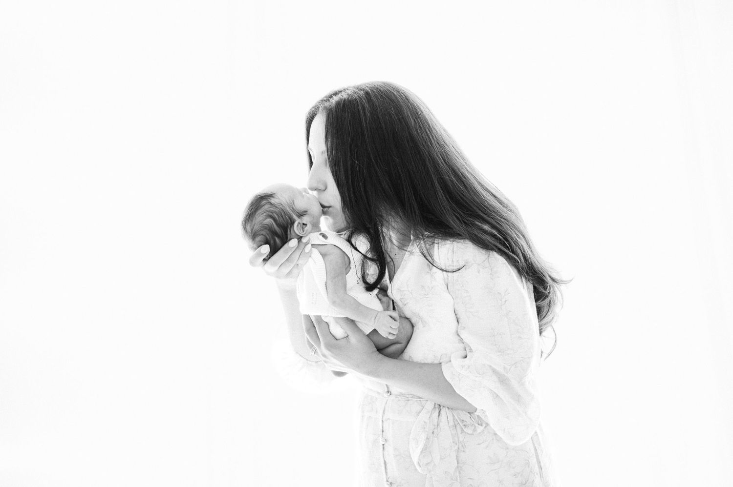 Black and white photo of a mother tenderly kissing her newborn baby against a bright white background.