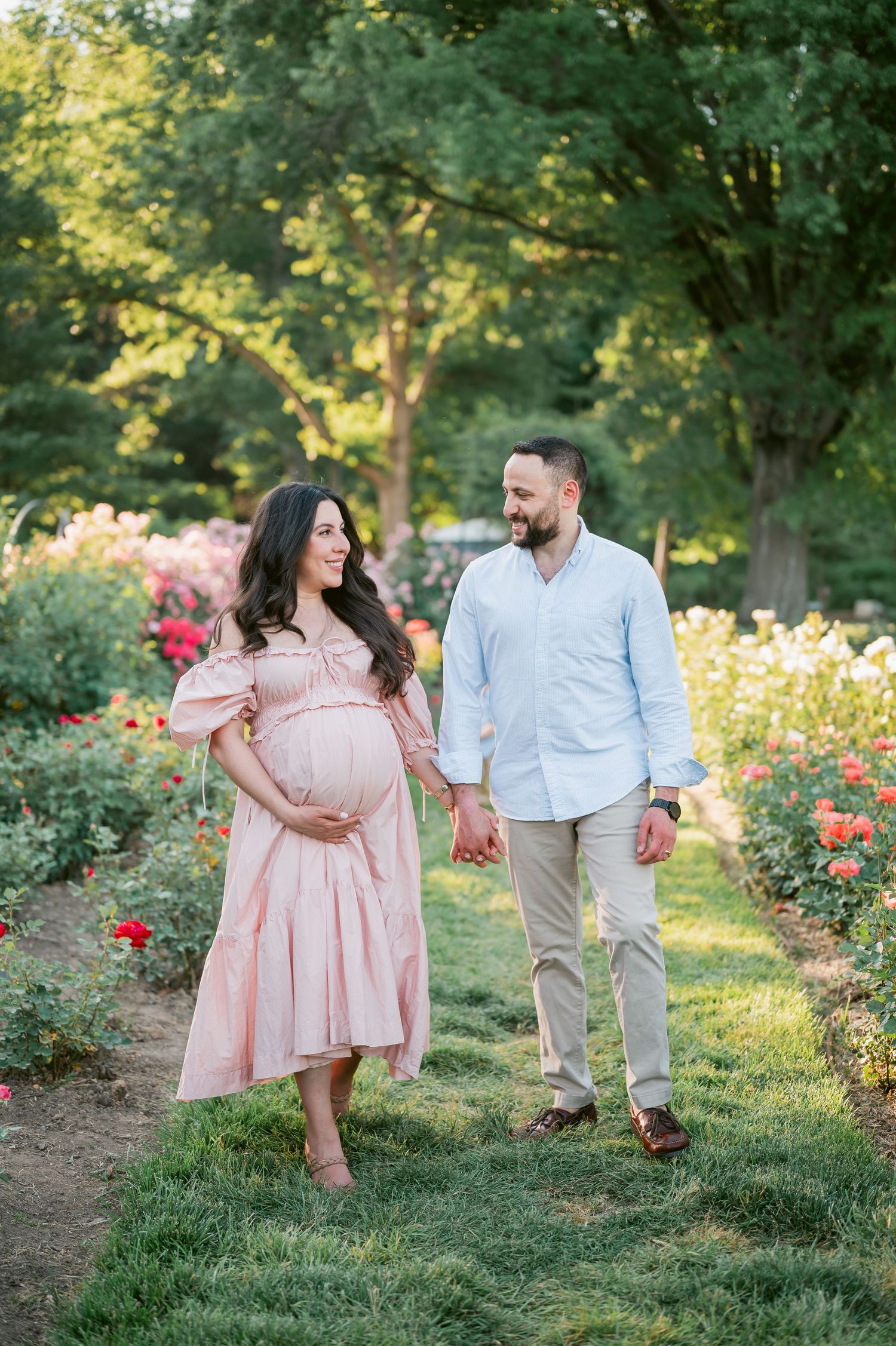 Pregnant couple holding hands walking through a blooming rose garden, woman in pink dress, man in light blue shirt.