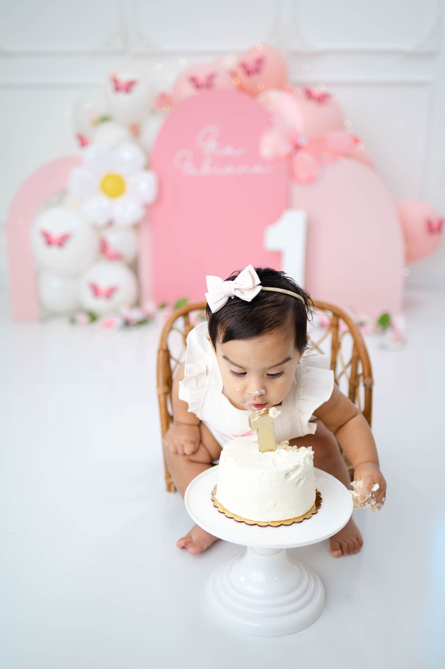 Pink butterfly and floral themed first birthday cake smash setup with white pedestal cake stand against pastel backdrop.