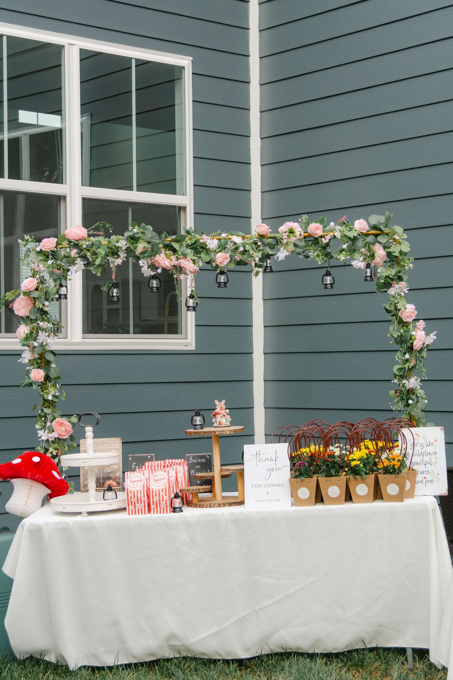 Long white buffet tables decorated with floral arches and party treats set up against a blue-gray house exterior.