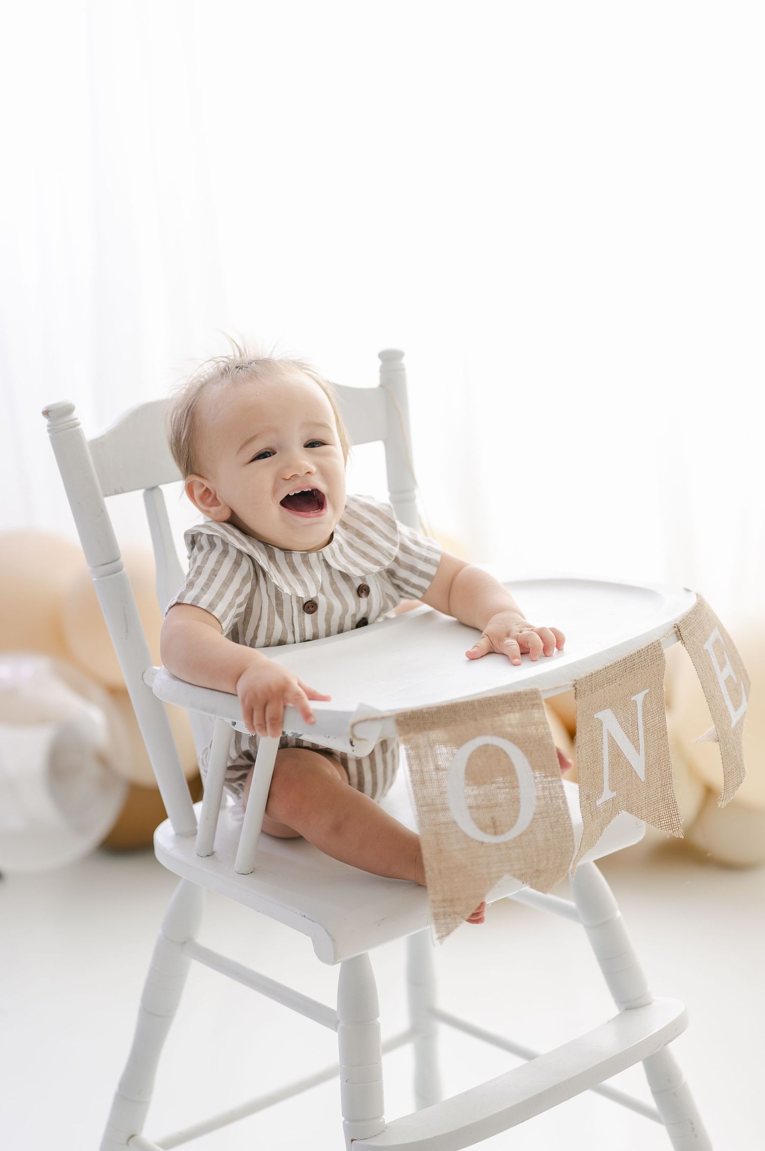 A laughing baby sits in a white high chair with a burlap ONE banner for their first birthday celebration.