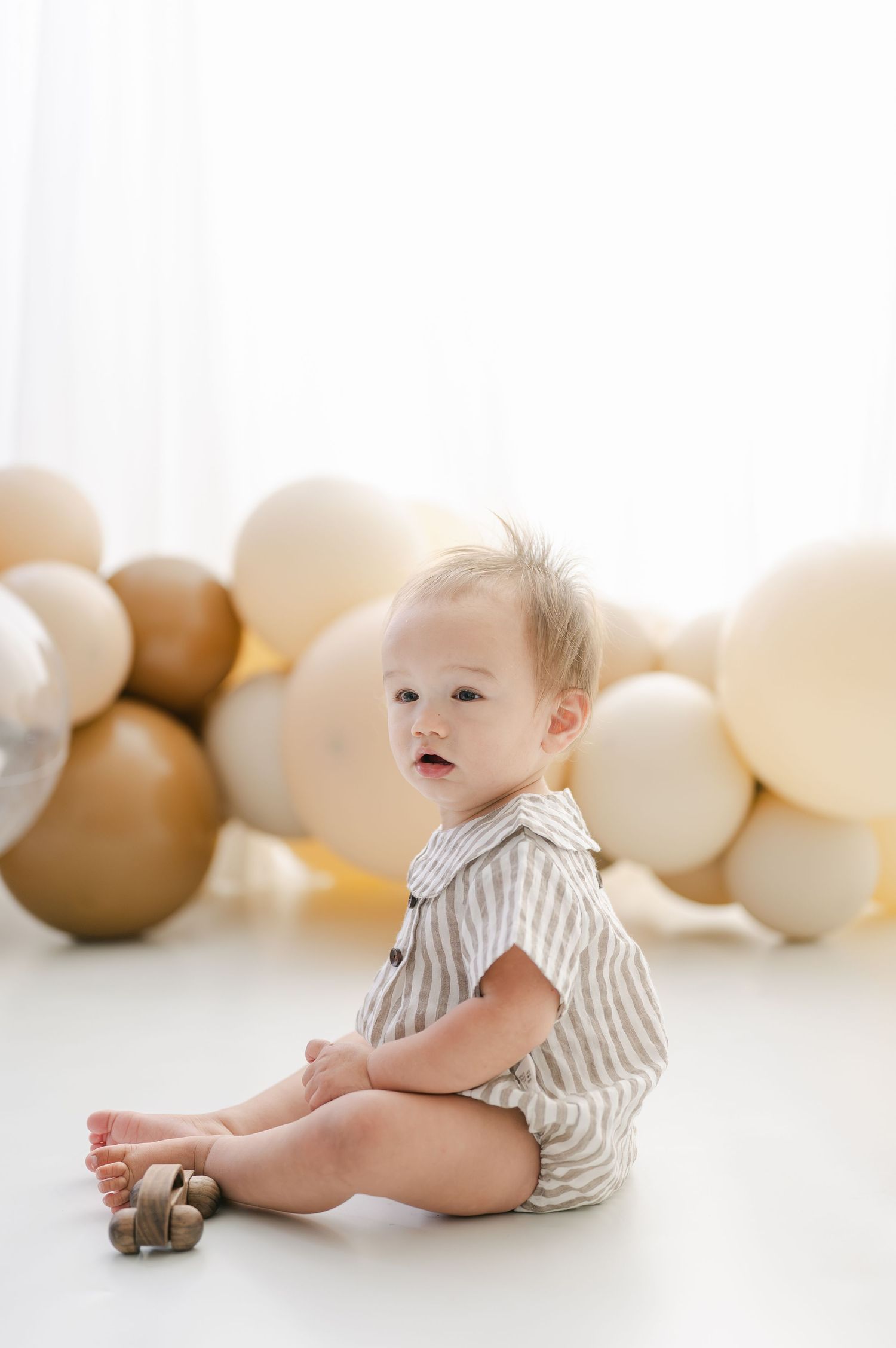 A baby boy in a striped romper sits on a white floor surrounded by neutral beige and brown balloons.