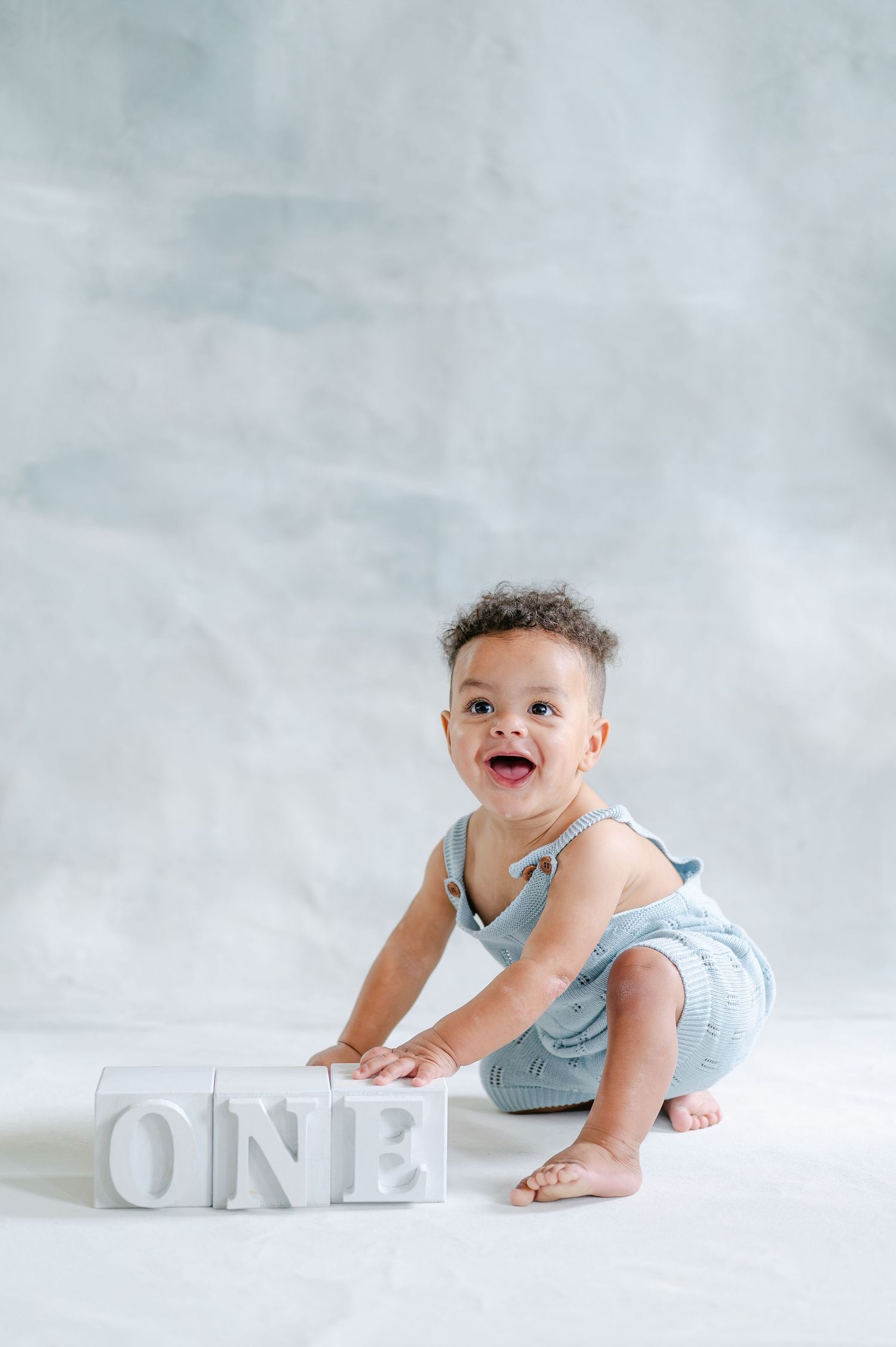 Smiling baby in blue romper crouching next to white letter blocks spelling ONE for first birthday photo.