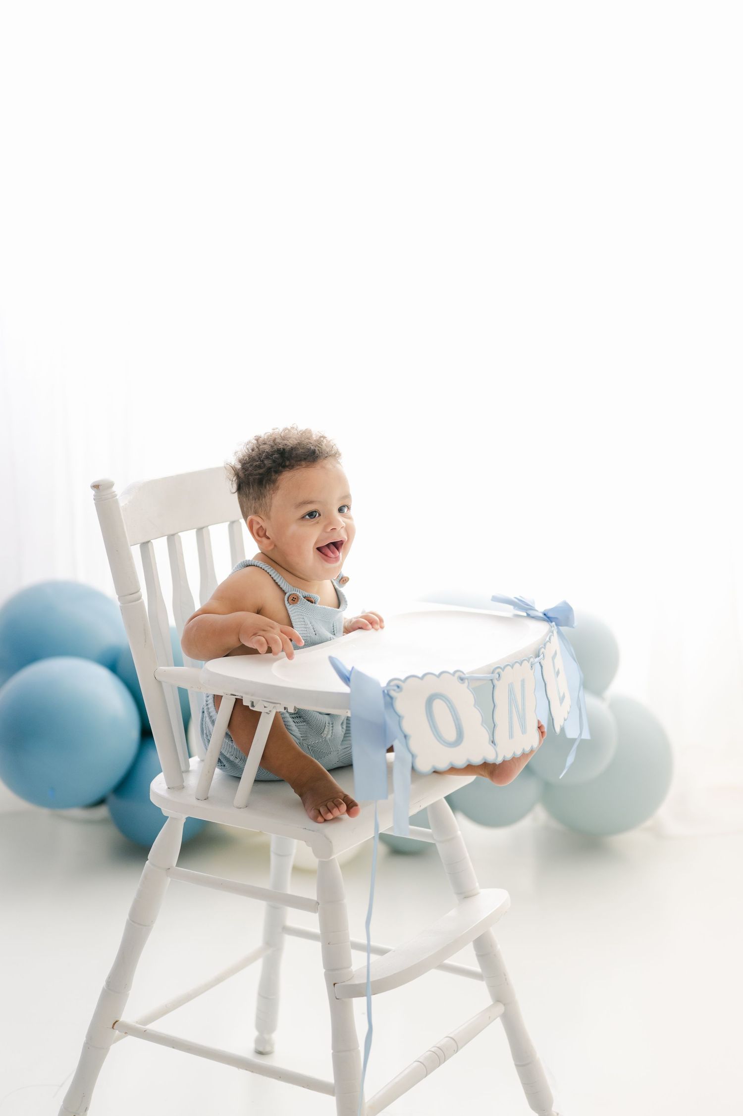 Happy baby in white high chair surrounded by blue balloons, celebrating first birthday with decorative banner.