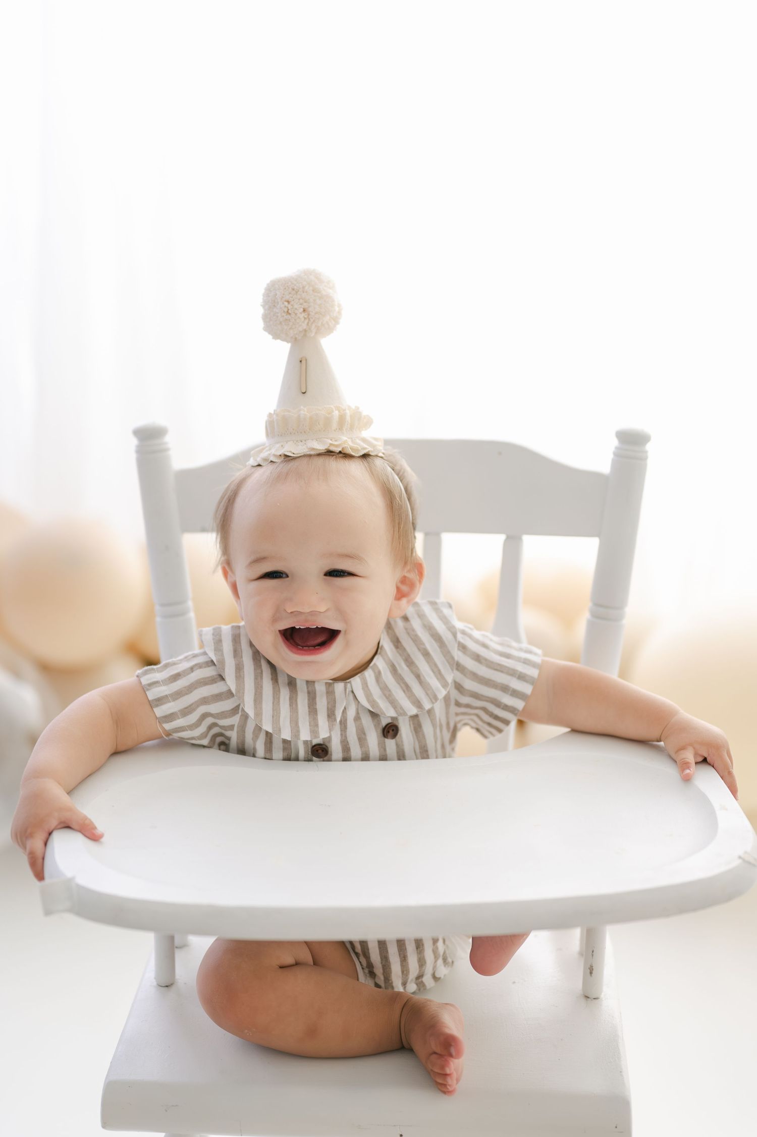 Smiling baby in striped romper and party hat sitting in white high chair, celebrating first birthday.