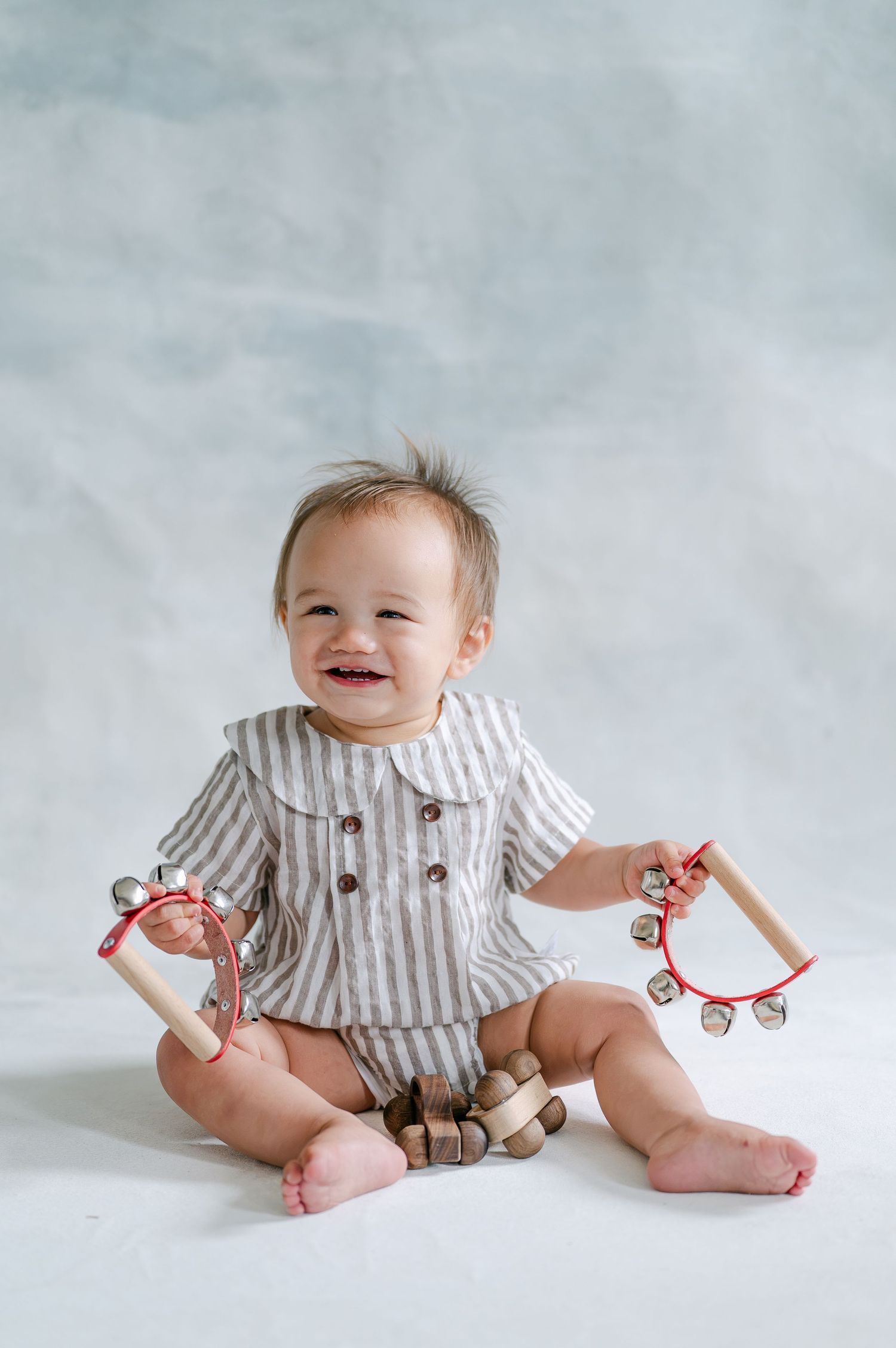 Smiling baby in striped outfit sitting on white background holding two small tambourines with red handles.